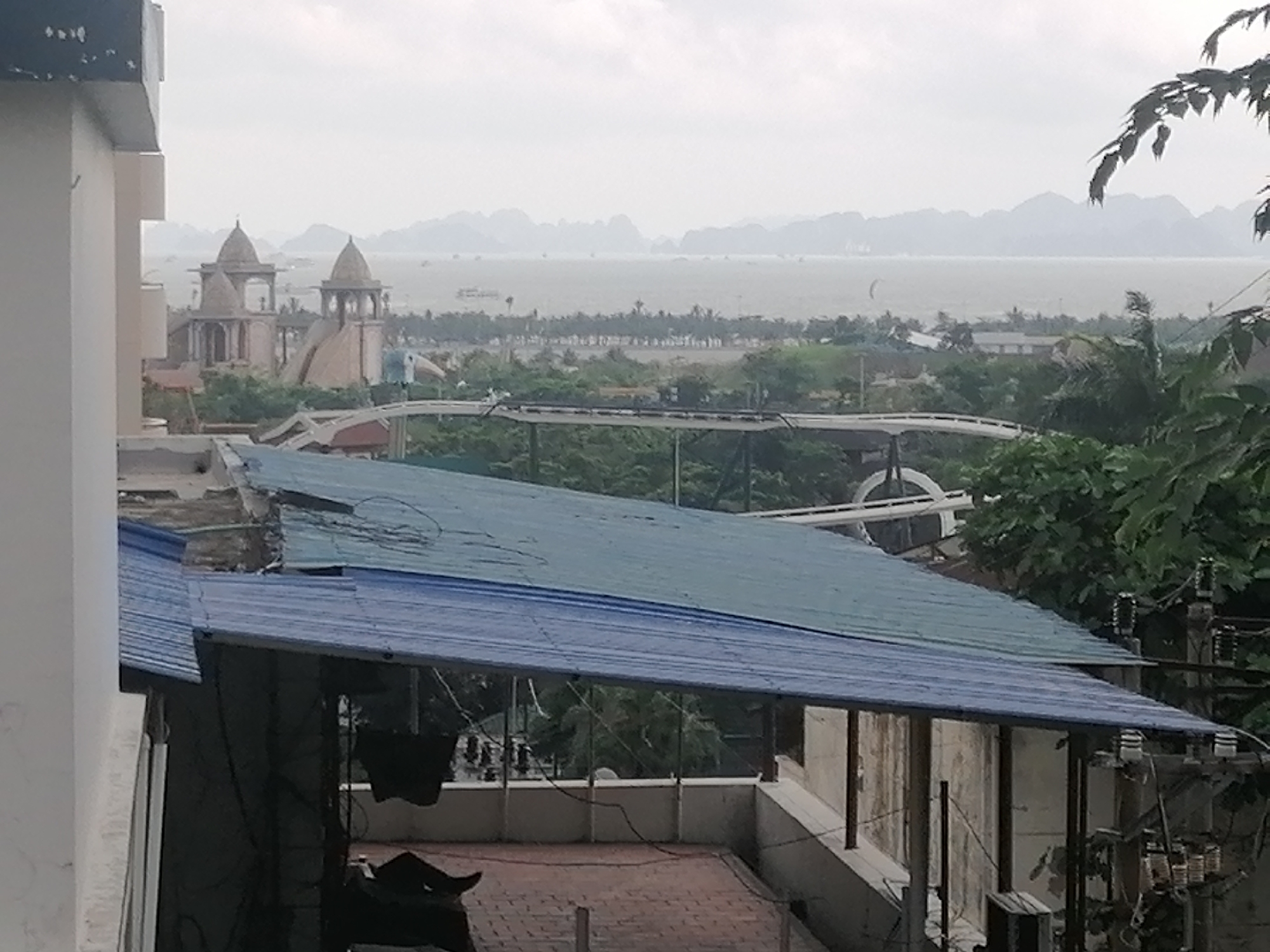 The image presents a panoramic view from an elevated position, looking out over a coastal area under an overcast sky.
In the immediate foreground on the left, a portion of a white building is visible. Below it, two blue corrugated metal roofs overlap, covering what appears to be a lower structure or terrace. On the terracotta-tiled terrace, partially obscured by the roofs and railings, a dark shape, possibly a person or an object, rests on a chair. Electrical wires and utility poles are prominent on the right side of the foreground.
The midground features an amusement park complex with distinct structures. A white rollercoaster track winds through abundant green foliage. Two prominent, ornate buildings with multiple domed roofs and arched windows, reminiscent of Indian or Middle Eastern architecture, stand out against the greenery. These light-colored structures appear to be part of a themed attraction. Further beyond, a strip of land with numerous trees, possibly palm trees, borders the water.
The background is dominated by a large body of water, likely a bay, appearing calm and grey under the hazy, overcast conditions. Several small boats are scattered across the water. In the far distance, numerous towering limestone karst mountains, characteristic of Ha Long Bay, rise from the water, their grey forms blending into the cloudy sky.
The overall scene depicts a developed coastal area with recreational facilities set against a dramatic natural landscape. The time of day is daytime, with diffused light indicating an overcast or cloudy weather condition. No discernible text is visible in the image.