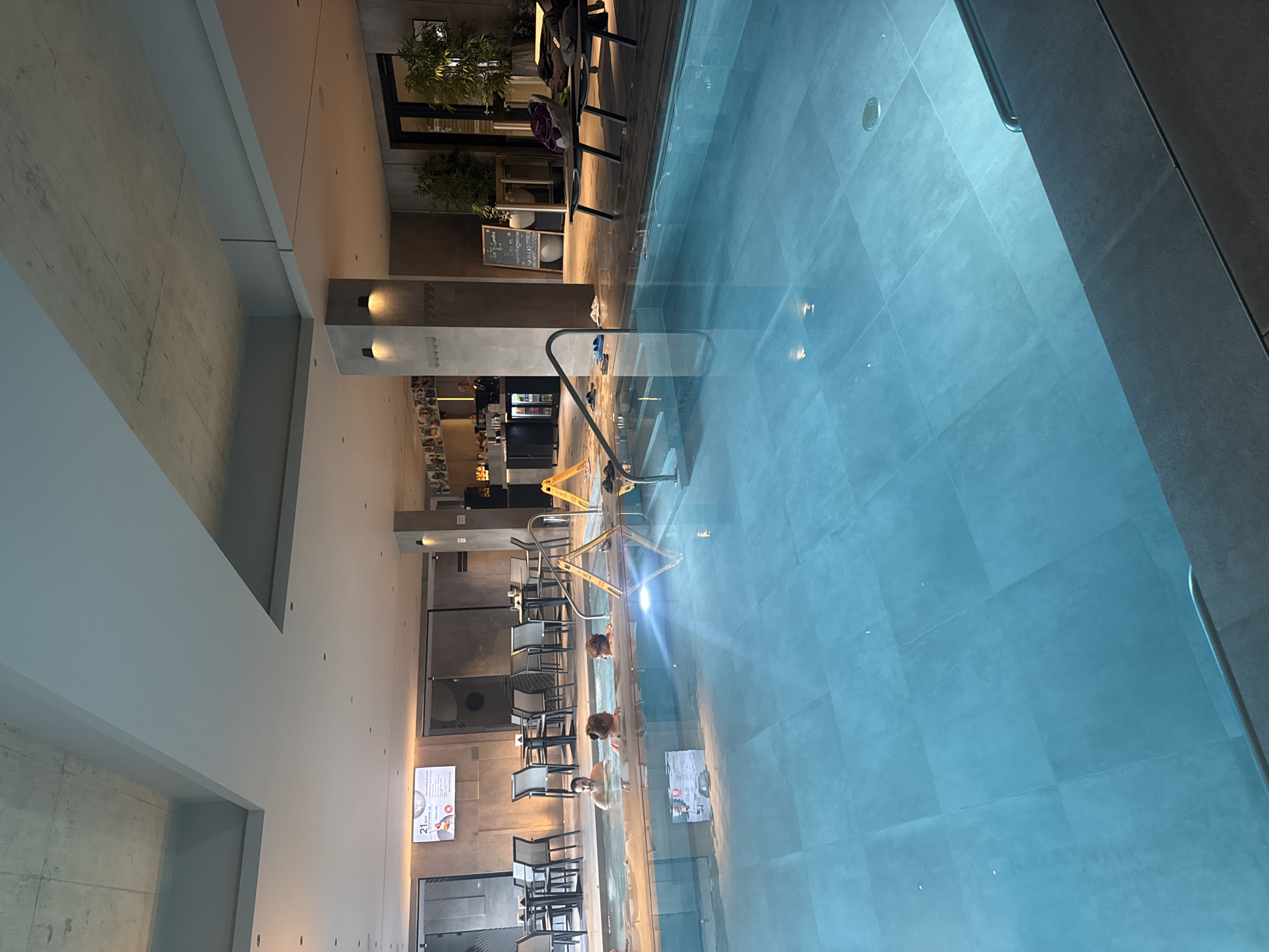 The image depicts an indoor spa and pool area within the Hotel Bulgaria Burgas, located at Aleksandrovska Street 21, Burgas, Bulgaria.
The scene features a large, rectangular swimming pool with blue-grey tiles in the foreground and midground, reflecting overhead lighting. To the left, a raised, smaller pool or hot tub is present, occupied by four individuals. One adult male, shirtless, is partially submerged, facing left. Two adult females are partially submerged, one facing left and the other facing right. A fourth, smaller individual is partially visible between the two females. All are stationary in the water. Metal handrails for entry/exit are visible on both pools.
The surrounding floor is tiled in a dark color and appears wet in areas. Several pairs of flip-flops are scattered near the pool edge. A yellow "WET FLOOR" sign stands near the left edge of the main pool.
In the background, a lounge area with multiple dark-colored tables and chairs is arranged to the left of the hot tub. A large digital display screen on the back wall shows "21" and text in Bulgarian. To the right, a fully stocked bar with bottles and glassware is visible, with a person's silhouette behind the counter. Further to the right, a dark chalkboard sign stands near two dark sun loungers on which towels and personal items are placed. Large potted plants are situated near a dark doorway in the far right background.
The ceiling features exposed concrete sections and suspended white panels, with recessed lighting throughout. Wall-mounted sconces provide additional illumination. Concrete support columns are visible, maintaining a modern, industrial aesthetic.