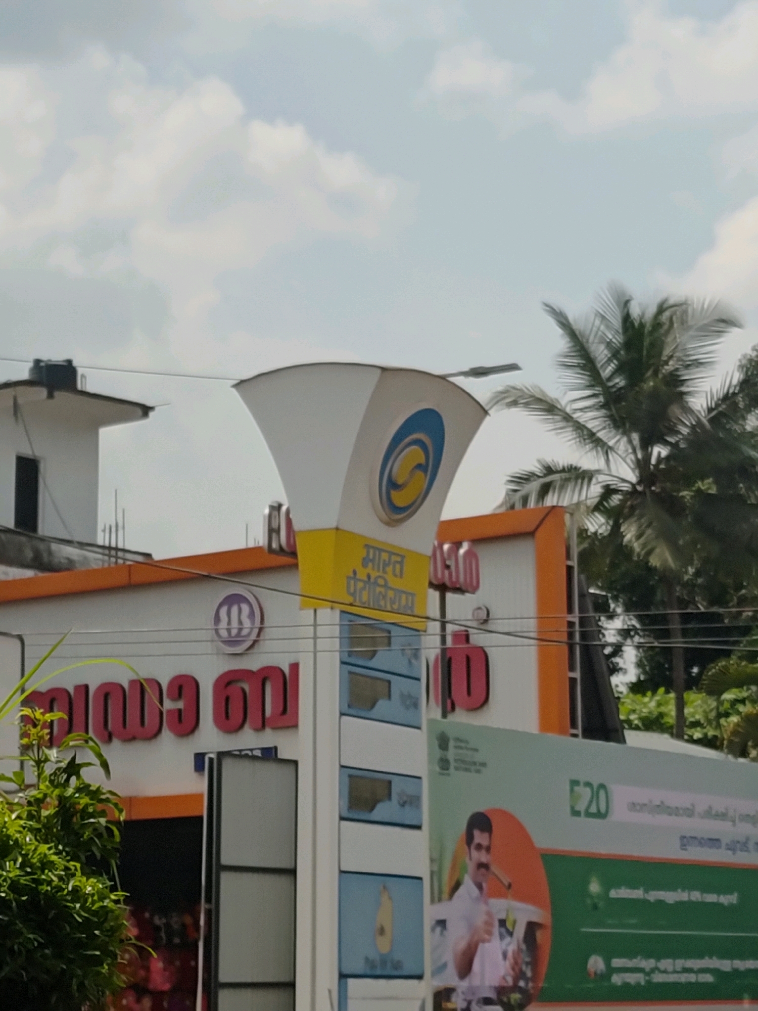 The image shows a Bharat Petroleum petrol station in Mannarkkad, India, under a cloudy sky with palm trees visible in the background. A large, white, conical structure with the Bharat Petroleum logo is prominent. Below it, a sign with red lettering in Malayalam reads "Raja B". A green banner on the right side of the image displays "E20" and a man giving a thumbs-up gesture, suggesting an ethanol blending initiative. Several other signs and structures are visible, including what appears to be a water tank on a building. The scene is typical of a roadside establishment in a tropical region.