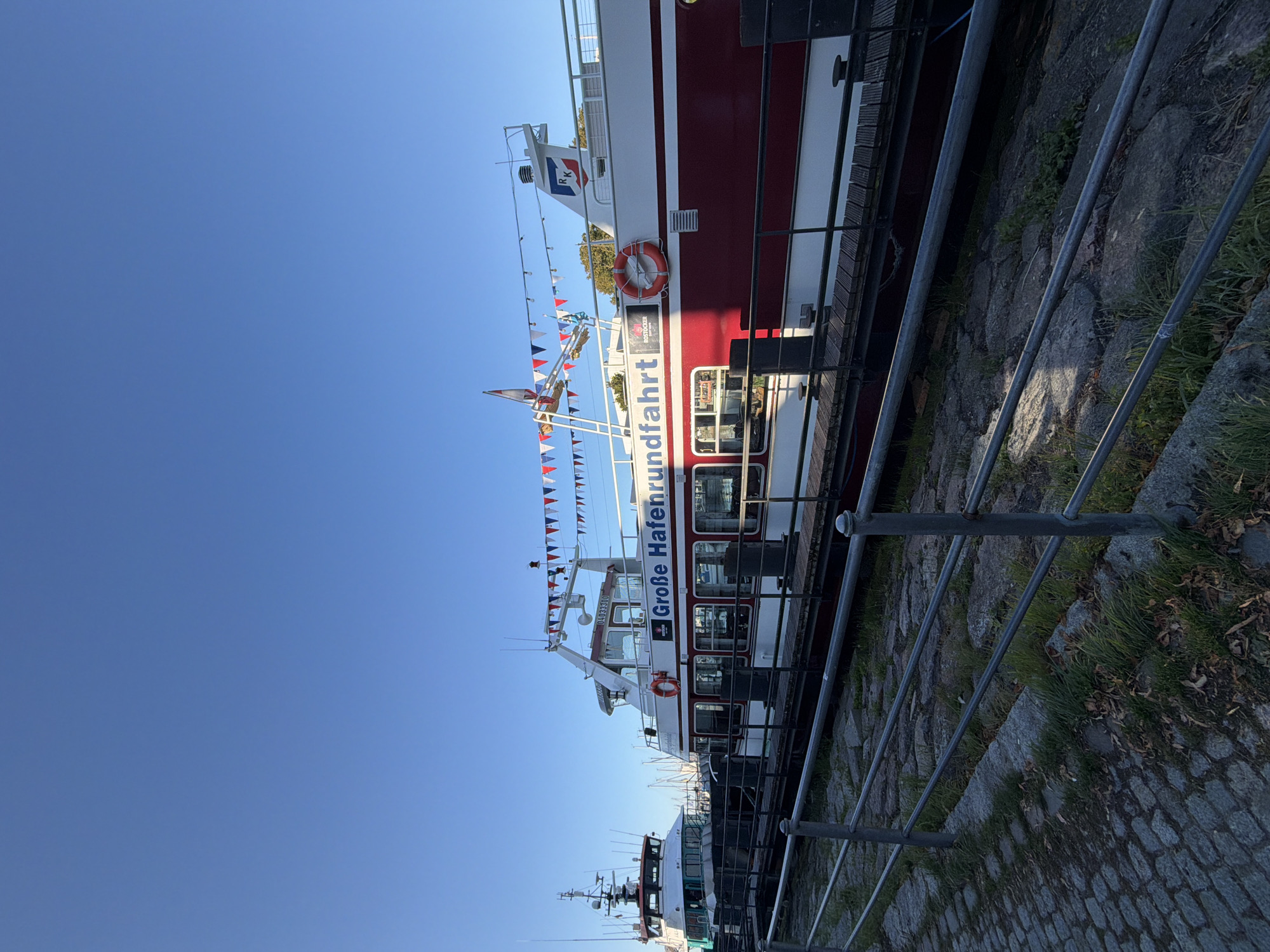 The image shows a bright, clear day with a blue sky. In the foreground, a cobblestone walkway lines a pier with a metal railing. Beyond the railing, two boats are docked. The boat on the right is larger and red and white, with "Große Hafenrundfahrt" (Great Harbor Tour) written on its side in large blue letters. It also has a life preserver and small triangular flags strung along its upper deck. The boat on the left is narrower and appears to be a working vessel with a white superstructure and green hull. In the background, more boats and masts are visible, suggesting this is a harbor or marina. The lighting indicates it is likely morning or late afternoon. The scene appears to be the Warnow River harbor in Rostock, Germany, specifically where harbor tours depart. There are no people visible in the image.