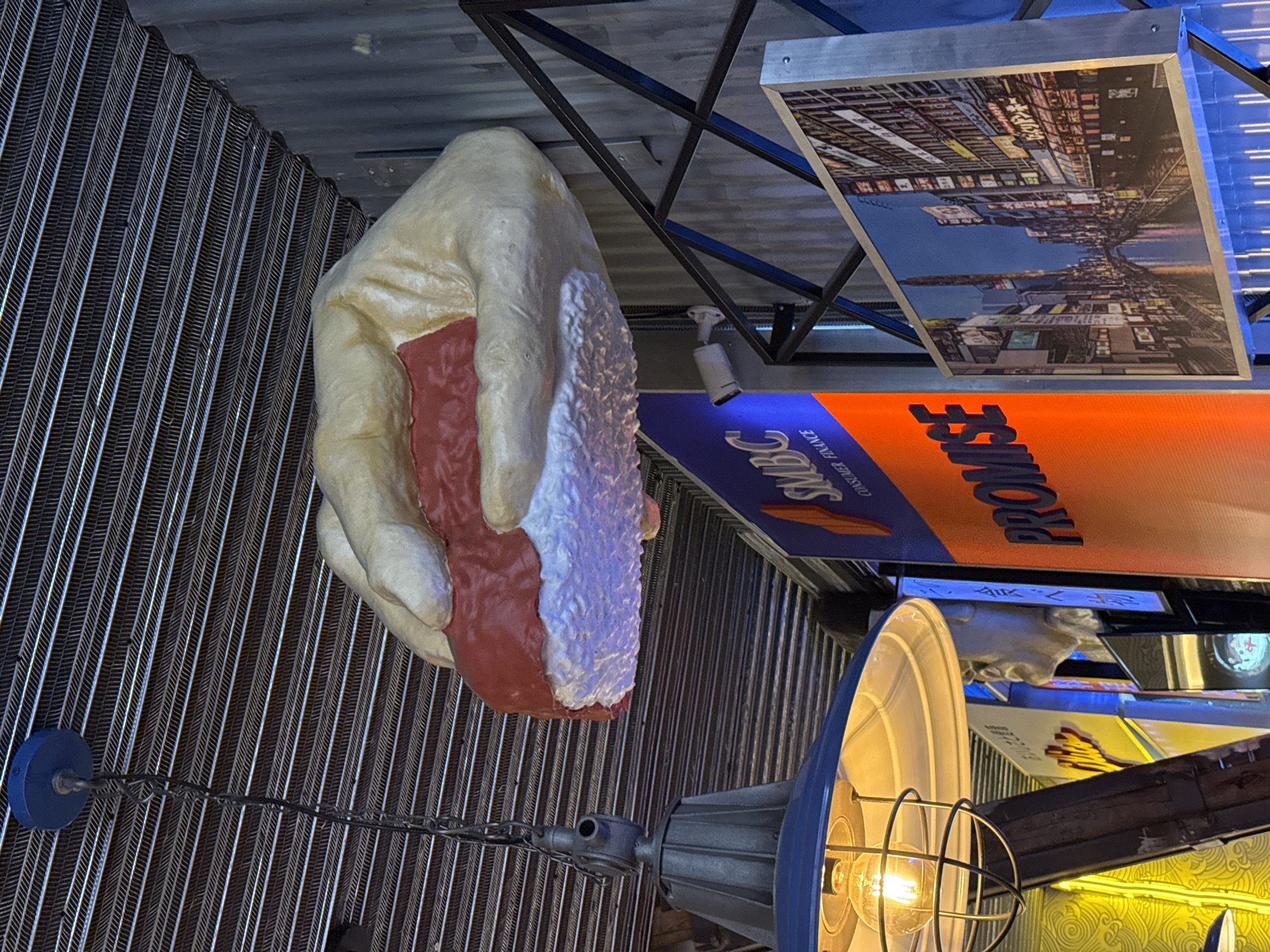 A view of an interior ceiling in Madrid, Spain. A large, oversized prop of a hand holding a slab of meat is suspended, along with signs and decorative lamps. One sign reads "Promse Consumer Finance" and another appears to be a photograph of a city street.