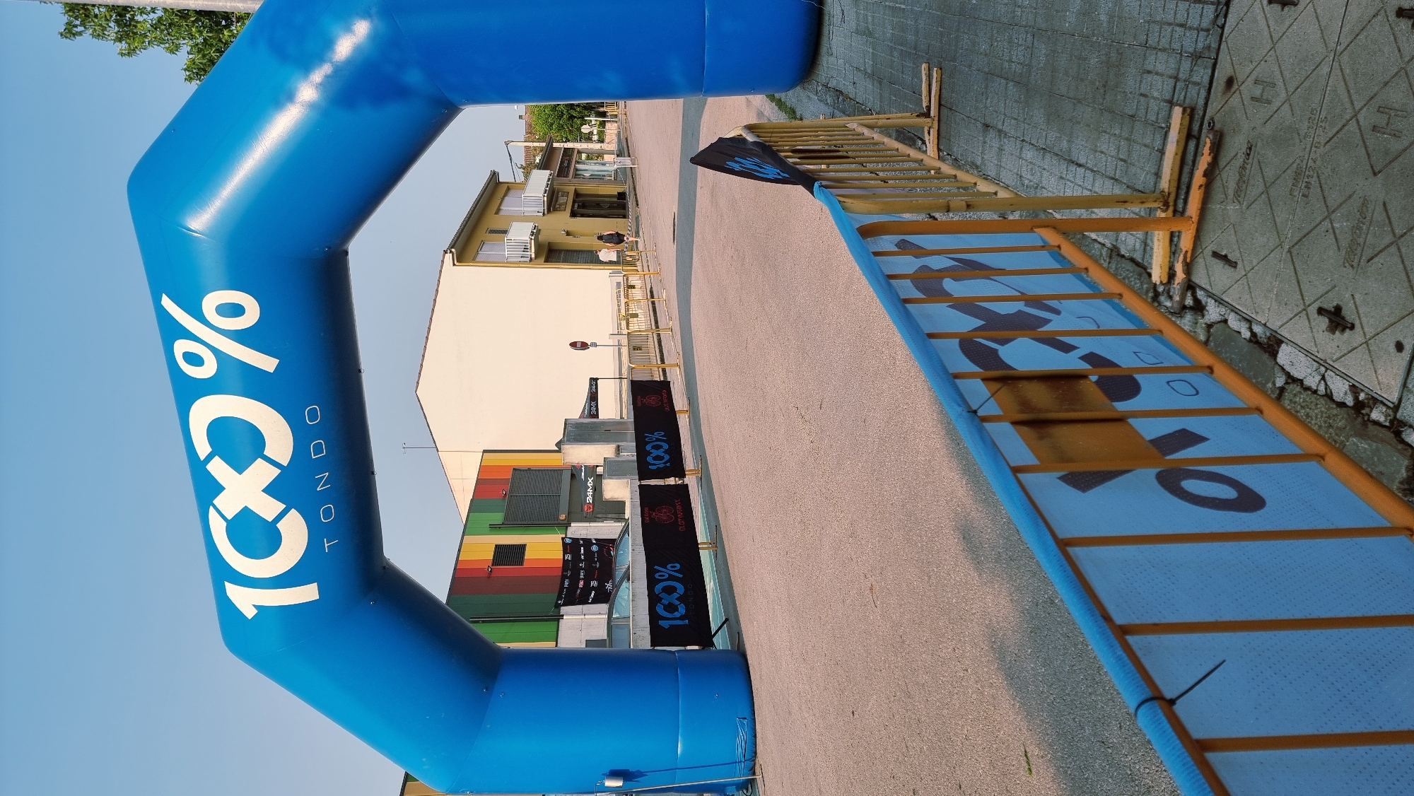 In the city of Sant Joan Les Fonts, Spain, an inflatable blue archway with white lettering reading "100% TONDO" stands prominently, marking the start or finish line of an event. Below the arch, black banners with "100%" and a logo are visible, attached to yellow safety barriers. A paved road stretches towards a building with colorful vertical panels. A sidewalk borders the road, also lined with yellow barriers. The sky above is clear and blue.