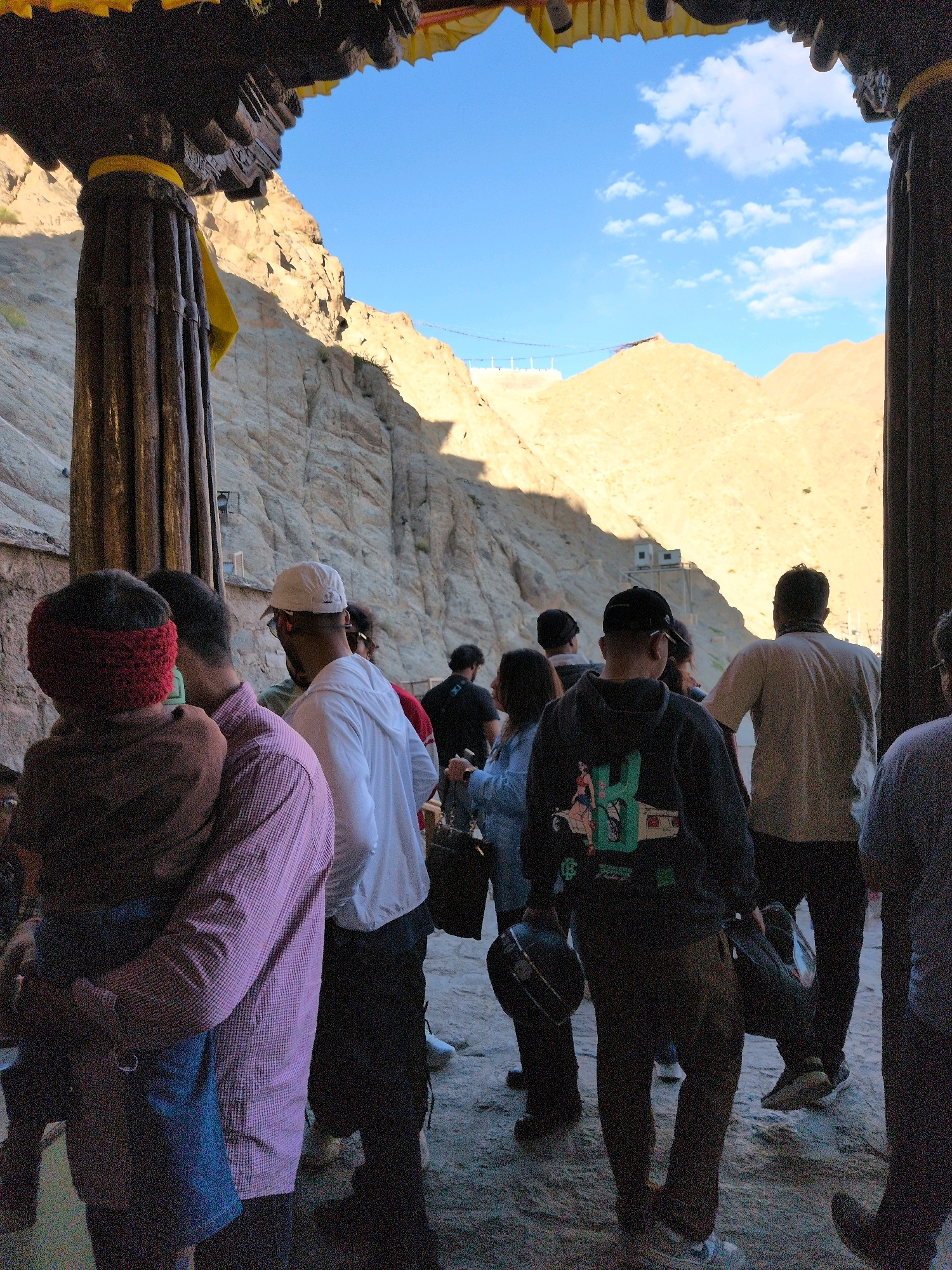 The image depicts a group of people standing at the entrance of a structure, possibly a monastery or temple, in the city of Leh, India. The entrance features large, ornate wooden pillars and a decorative archway with yellow fabric. Beyond the entrance, a vast, arid landscape with sun-drenched rocky hills and a bright blue sky with scattered clouds is visible.
In the foreground, several individuals are positioned, some facing away from the camera and others partially obscured. One person in a red headband is holding a child. Other individuals are dressed in casual attire, including a person in a pink checkered shirt, someone in a white hooded garment, and another in a black hoodie with graphics. Some are carrying bags or helmets. The ground appears to be sandy or dusty. The overall scene suggests a tourist or pilgrimage site, with visitors preparing to enter or having just exited the structure.