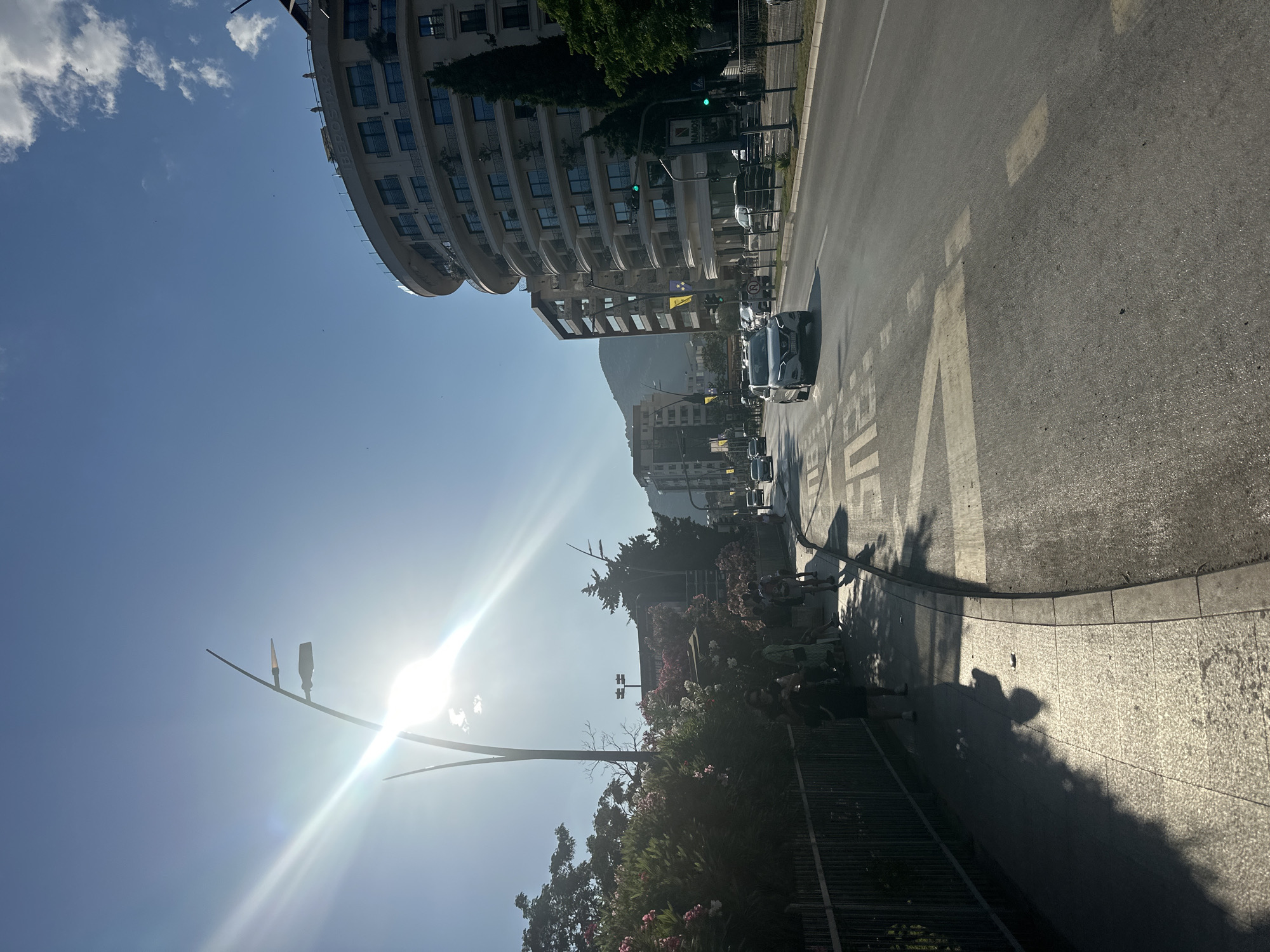 This image depicts a street scene in Budva, Montenegro, on a bright, sunny day. The viewpoint is from a sidewalk looking out onto a road. The sun is high in the sky, casting strong shadows and creating lens flare.
On the left side of the image, there is a sidewalk with people walking and sitting. Some are silhouetted against the bright sunlight. There are lush green bushes with pink and white flowers along a fence separating the sidewalk from the road. A modern street lamp with multiple lights stands tall.
The road is paved and appears to be a main thoroughfare with multiple lanes. Several cars are visible on the road, including a white SUV in the foreground. Road markings, including a "BUS" designation, are clearly visible.
In the background, there are several multi-story buildings. A prominent, curved, cream-colored building with balconies dominates the right side of the frame. Beyond that, other urban structures and a green-covered hill can be seen under a clear blue sky with scattered white clouds. The overall atmosphere suggests a busy urban environment during daylight hours.