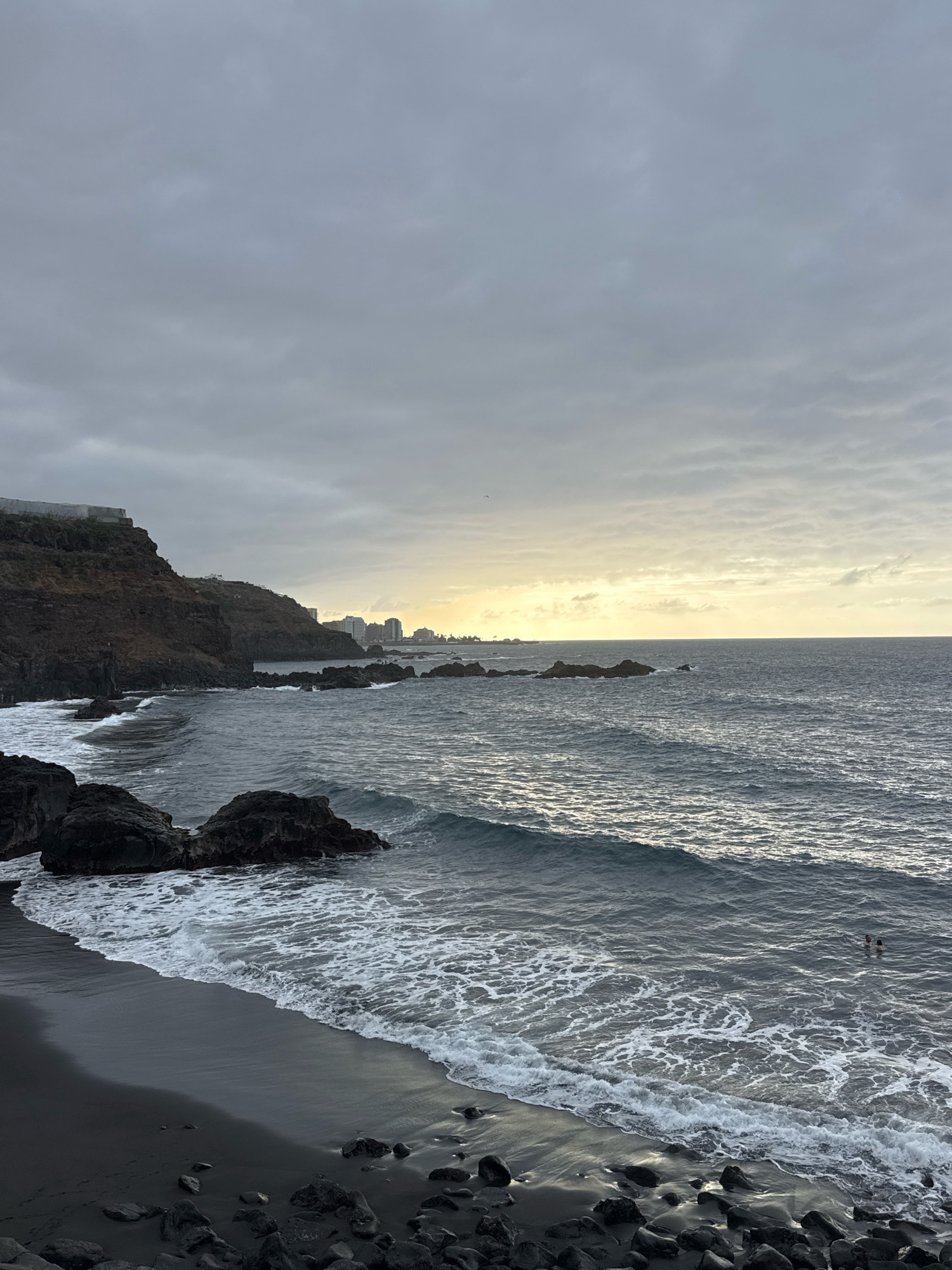 The image depicts a coastal scene with a black sand beach on the left and a calm ocean extending to the right. Jagged dark rocks are scattered along the shoreline and break the water's surface further out. In the distance, a city skyline with several tall buildings is visible along the coast. The sky is overcast with a soft, diffused light suggesting dawn or dusk. Two people are swimming in the ocean on the right side of the image. The overall mood is serene and natural.