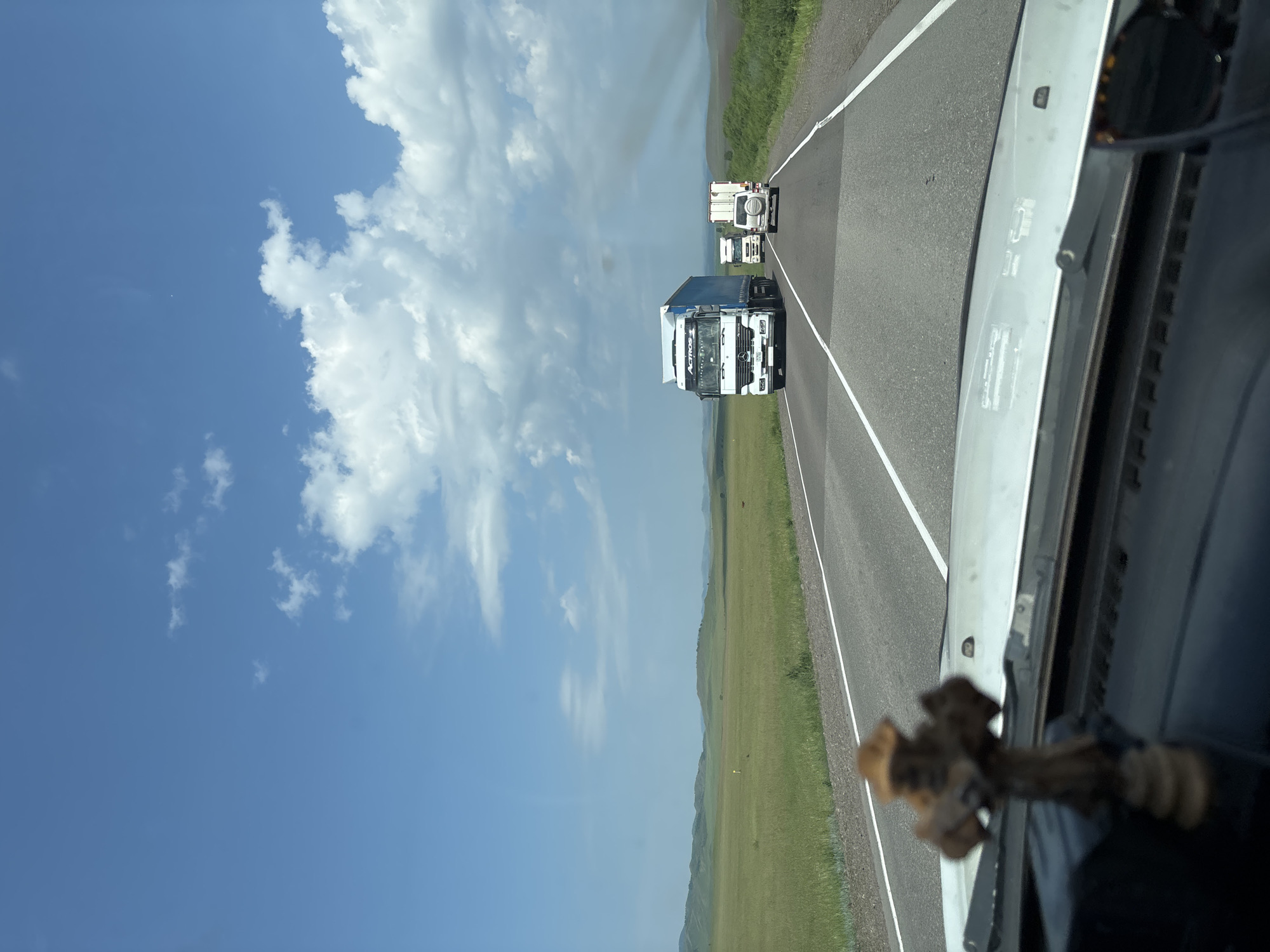 The image shows a view from the dashboard of a vehicle driving on a highway in Borodino, Russia. The road has multiple lanes and is bordered by grassy fields and rolling hills. In the distance, several trucks are traveling on the same highway. The sky is blue with scattered white clouds. A small wooden cross is visible on the dashboard of the vehicle in the foreground.