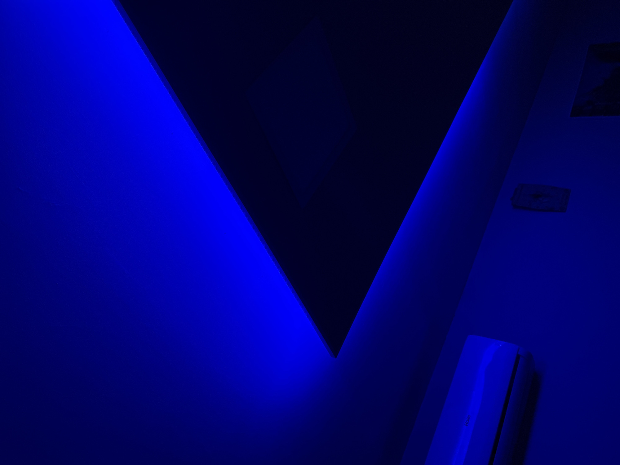 The image shows an interior scene illuminated by blue light. In the lower left corner, a white air conditioning unit is mounted on a blue wall. Above the air conditioner, the ceiling transitions from a bright blue to a darker blue and then to black. A thin strip of blue light runs along the edge where the ceiling meets the wall. To the right of the air conditioner, a small, square decorative object with a textured surface is mounted on the wall. Further to the right and lower on the wall, a framed picture is visible, but its contents are obscured by the low light. The overall environment appears to be a room with modern, minimalist design elements.