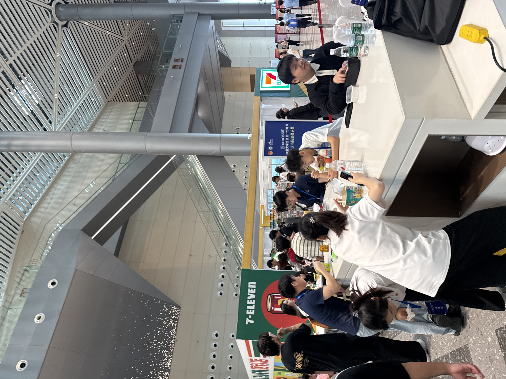 The image depicts a busy indoor environment, likely a convention center or exhibition hall, with a prominent 7-Eleven booth. Several people are seen gathered around a counter, with some appearing to eat or drink. The overall atmosphere suggests an event or public gathering. The presence of a 7-Eleven booth indicates a commercial or promotional activity. The architectural style, with large pillars and a modern ceiling, suggests a contemporary public space. The background shows escalators and signage, further reinforcing the impression of a large public facility. The people are dressed in casual attire, and their interactions appear to be social or transactional, typical of such an environment. There is a general sense of activity and movement within the space. The image is shot from a medium distance, capturing a wide view of the scene.
