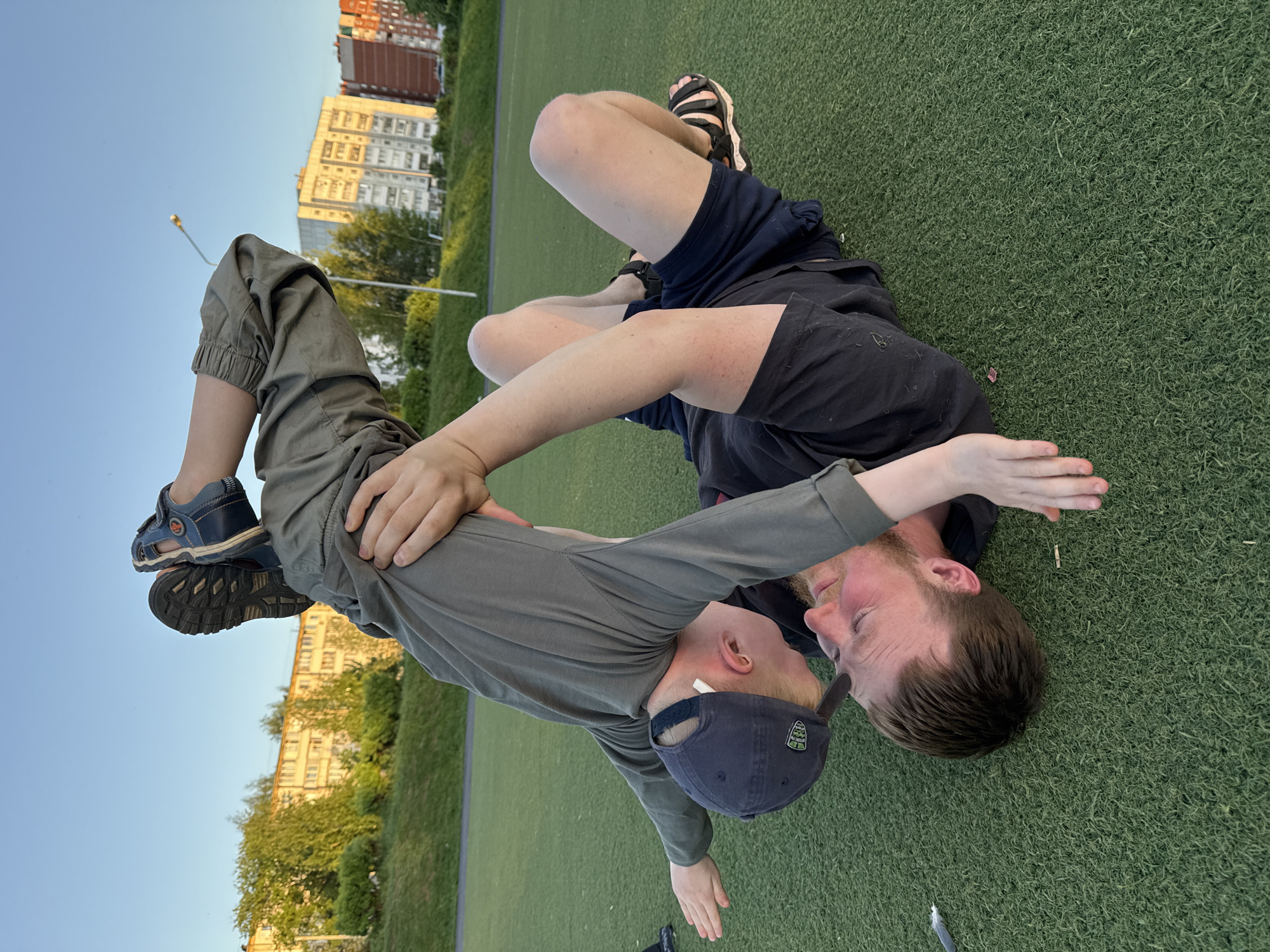 On a green artificial turf field in Chaikovsky, Russia, a fair-skinned man with reddish hair lies on his back. He wears a black t-shirt and dark shorts. A younger person with fair skin, wearing a long-sleeved gray shirt, dark green pants, and a blue baseball cap is positioned atop the man, facing away from the camera. The younger person's feet are in the air, held by the man. Buildings and trees are visible in the background under a clear sky.