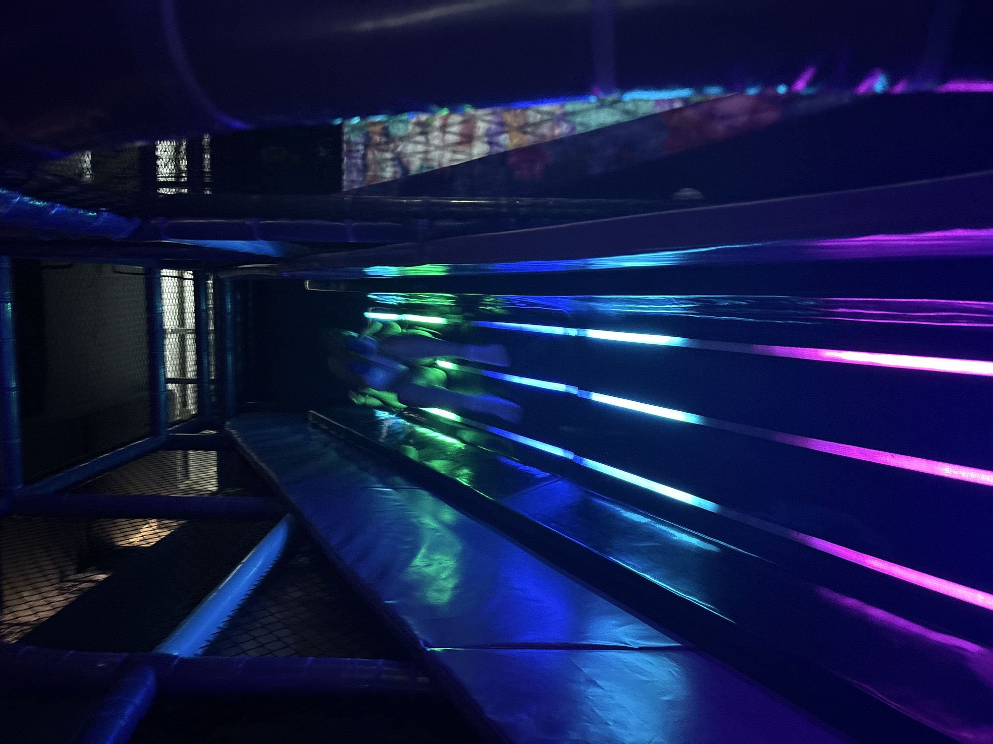The image depicts an indoor playground structure with a vibrant, futuristic aesthetic likely taken in an entertainment center. The environment is dark with artificial lighting creating a neon-like effect. The structure is a series of platforms or walkways constructed with padded surfaces and netting for safety. Dominant colors are blues, purples, and greens from light sources, possibly LED strips or other lighting fixtures. There is a sense of depth with elements extending into the distance, including what appears to be a window or opening with netting. The location in Izhevsk, Russia cannot be determined based on the image. No people are clearly visible in the image.