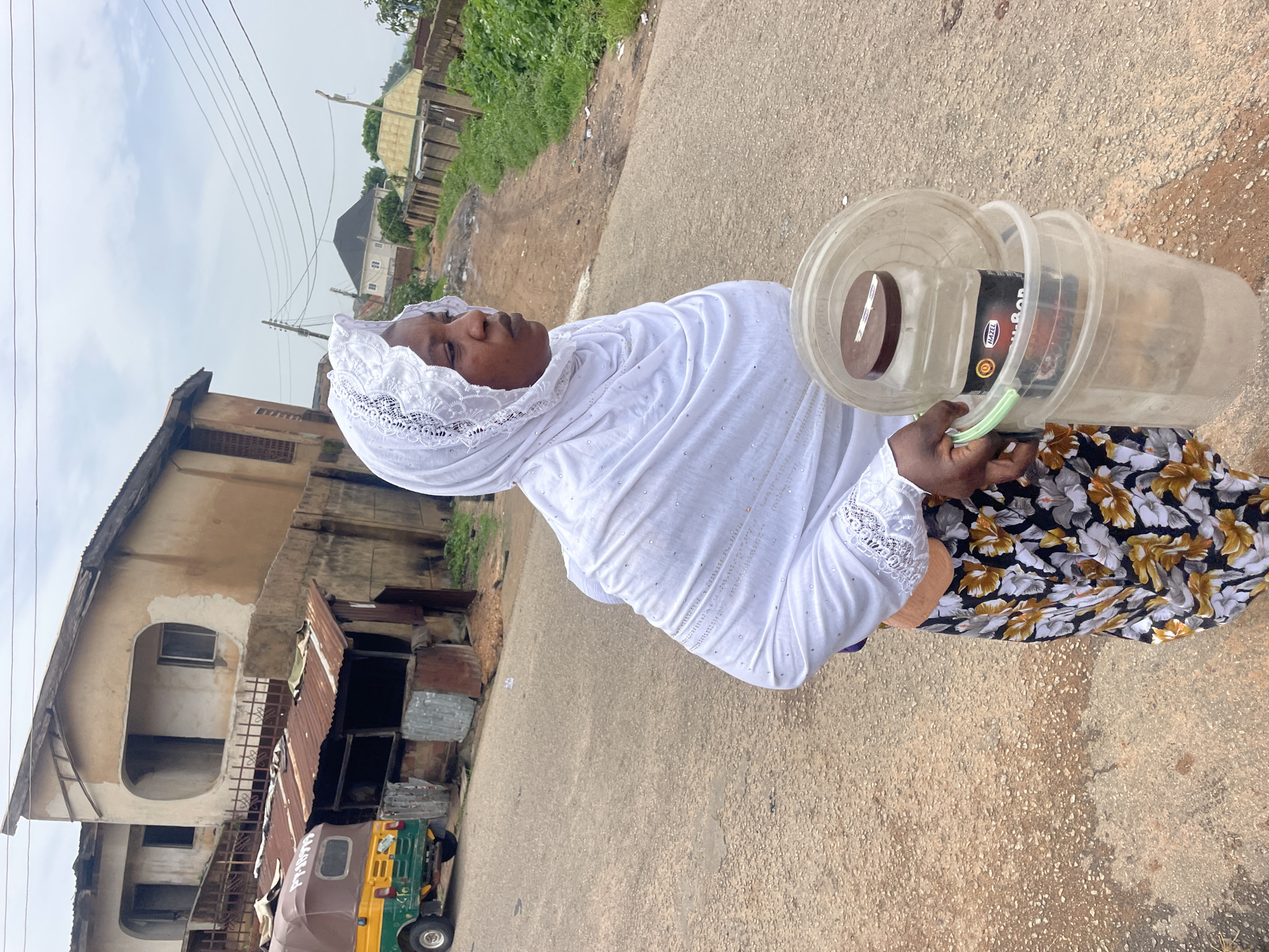 A person is standing on a street, holding two clear plastic buckets. The person is wearing a white headscarf and a floral skirt. The person's face is partially visible, and they appear to be looking down. One of the buckets contains a dark-colored item with text. The scene is outdoors, with buildings and vegetation visible in the background. The ground is paved with asphalt. A three-wheeled vehicle is parked nearby. The sky is overcast, suggesting it might be daytime, possibly with cloudy weather. The overall setting appears to be a residential or commercial area. The text on the item in the bucket is partially legible, appearing to be "HAZEL N-BODY".