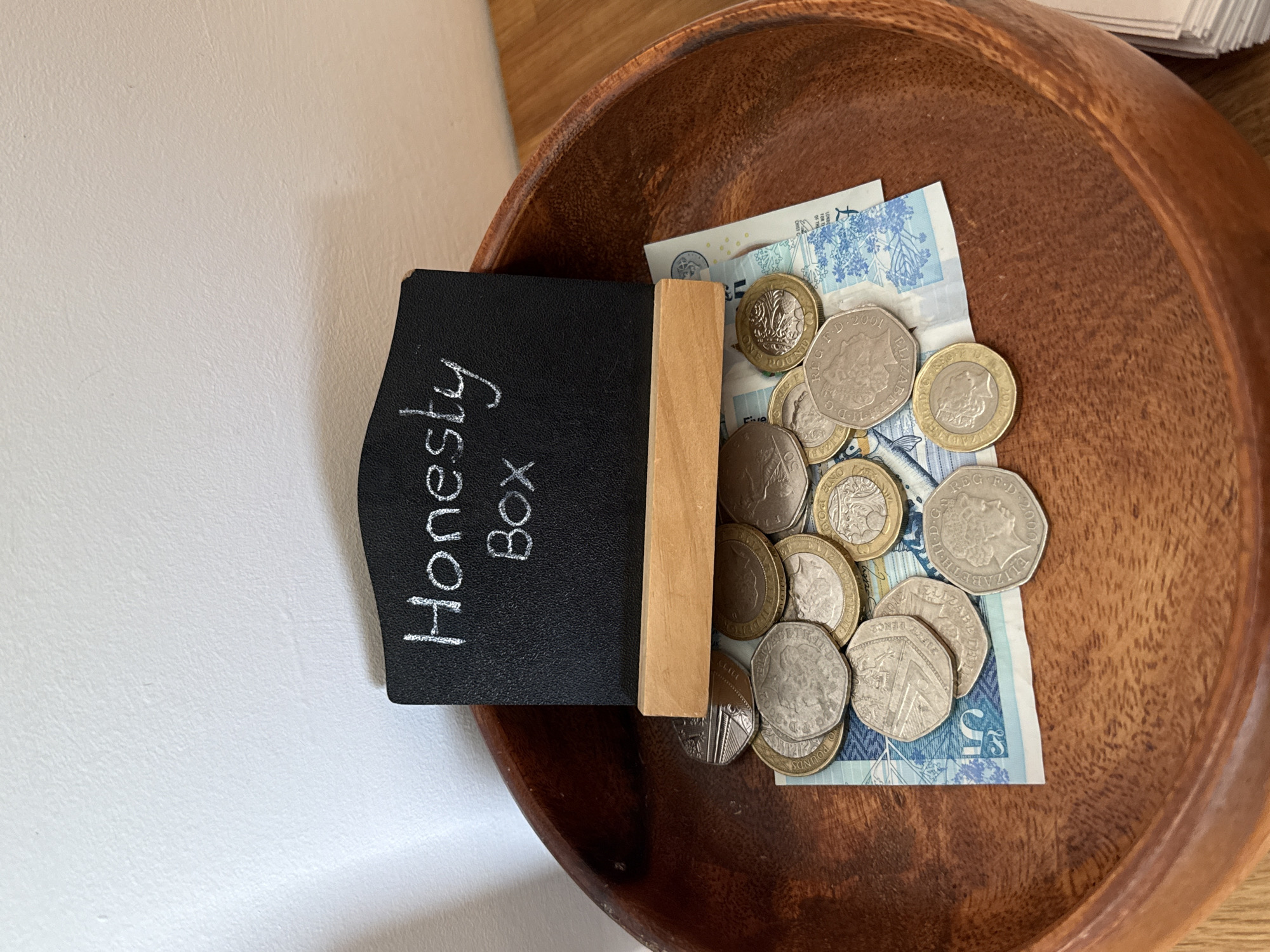 The image shows a brown wooden bowl containing UK currency (coins and a £5 note), and a small chalkboard sign that reads "Honesty Box." The setting appears to be indoors, with a white wall on the left and a light-colored wooden surface in the upper portion of the frame. No people are visible. The location cannot be determined.