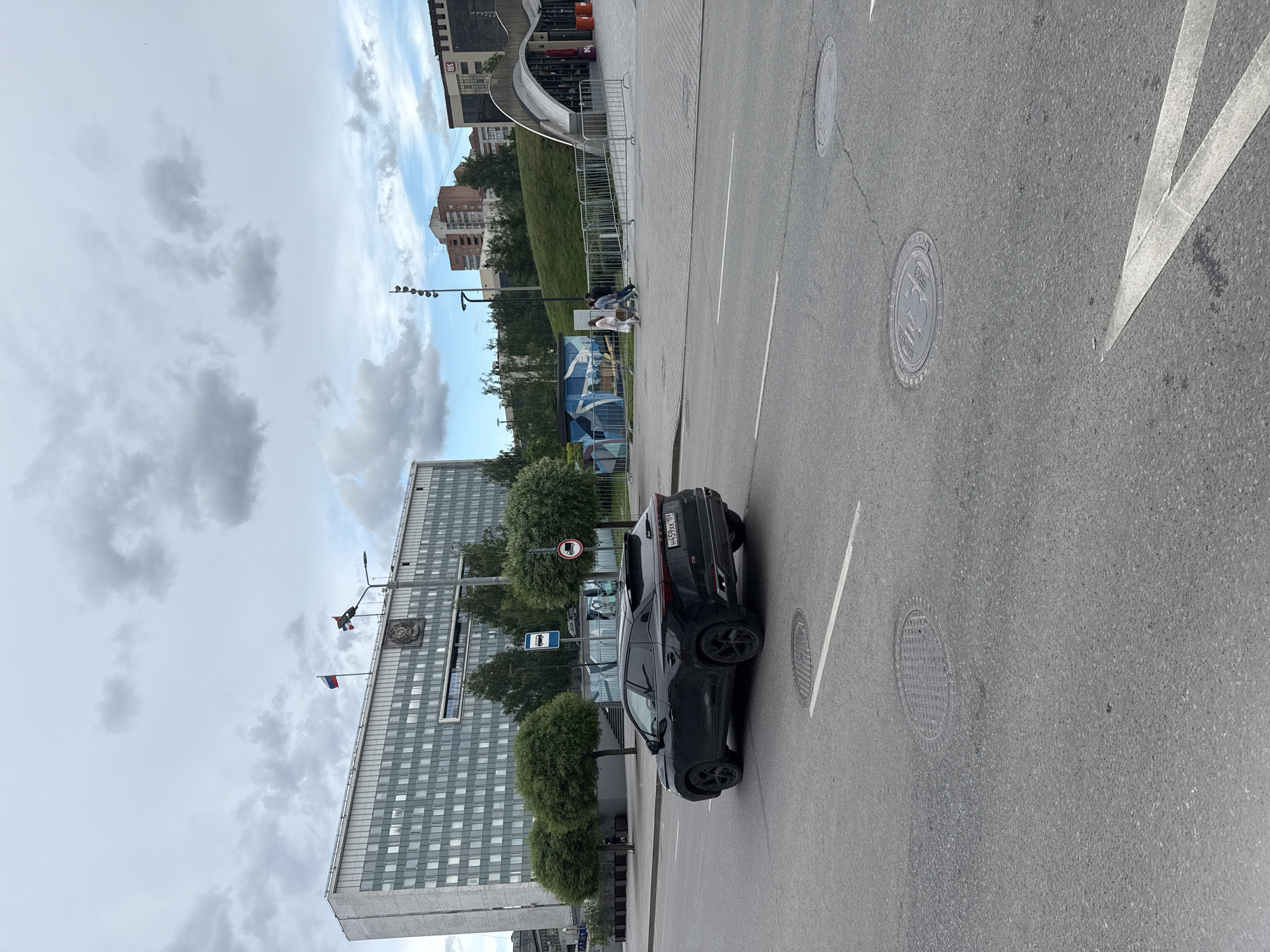 In the city of Perm, Russia, a black Chery Tiggo 7 Pro is driving on a paved road under a cloudy sky. The car is traveling away from the viewer, and its license plate reads H459KK159. To the left of the road is a large, grey, multi-story building with many windows, flying two Russian flags on its roof and a large emblem. In front of the building, there are several round, green trees. To the right of the road, there is a sidewalk with a metal railing, a bus stop, and a mural. Further in the background, there are more buildings and a grassy hill. Three people are walking on the sidewalk in the distance. The road has multiple manhole covers and white lane markings.