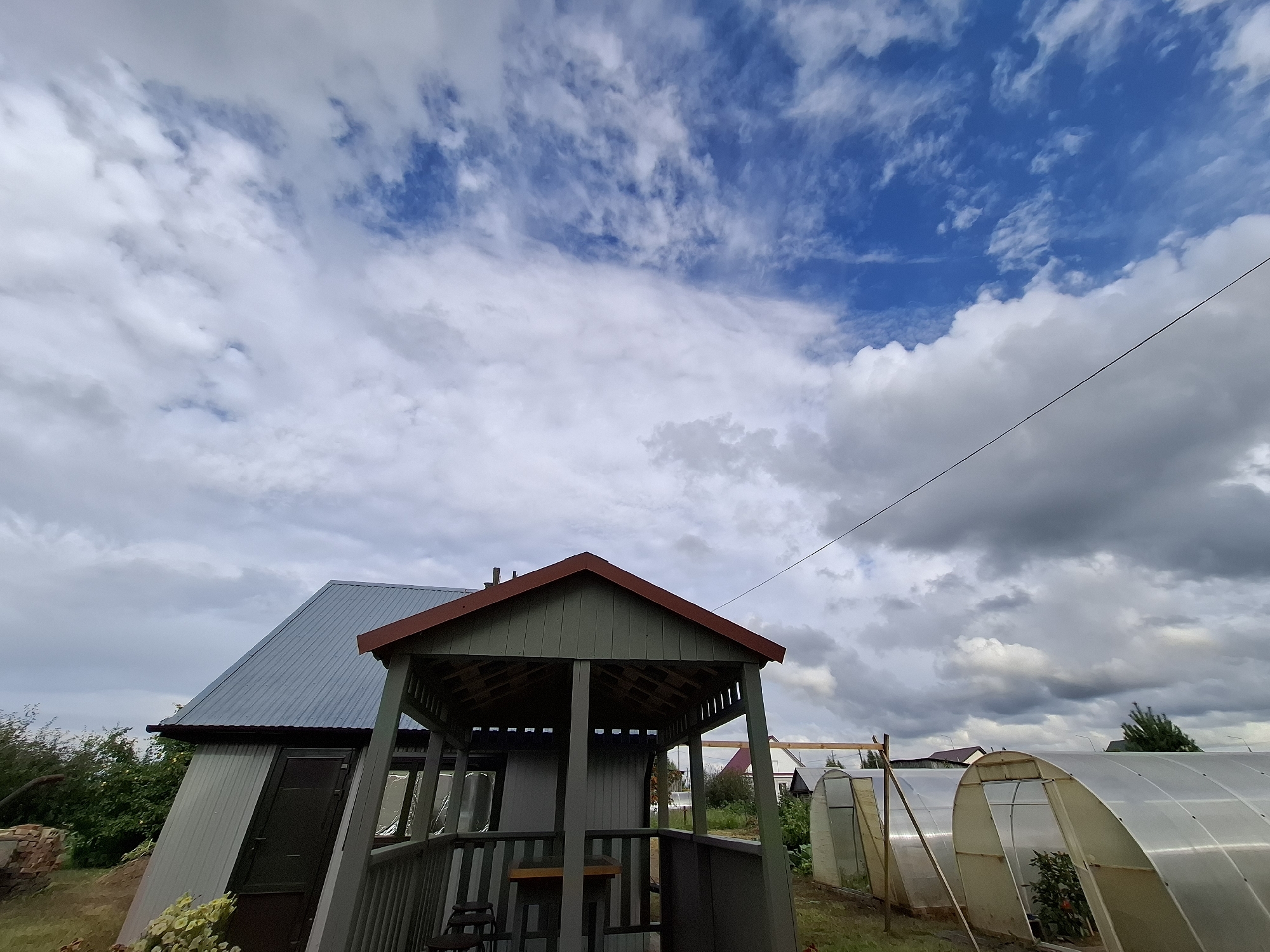 The image depicts a rural outdoor scene on a cloudy day. In the foreground, a green, open-air gazebo with a reddish-brown roof stands prominently. To the left of the gazebo is a gray building with a sloped metal roof and a dark door. To the right of the gazebo, two arched greenhouses made of transparent material are visible. The background shows some trees, a few houses with rooftops, and a utility wire stretching across the sky. The overall setting suggests a residential property, likely a dacha or a private home in a village. The location is identified as Амзя, Russia. There are no people or animals present in the image, and no actions or interactions are taking place. The environment is characterized by a mix of constructed elements and vegetation under a dynamic sky with both bright blue patches and heavy clouds.
