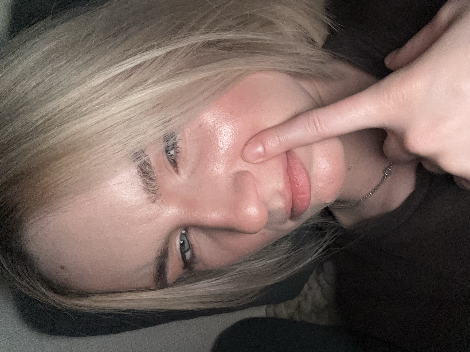 A close-up, tilted selfie shows a person with fair skin and light blonde hair lying down. The person is touching their face with their right index finger, pressing on their cheek near their lips. They have blue eyes and visible eyebrows. A silver chain necklace is visible around their neck, and they are wearing a dark-colored shirt. The background appears to be a dark-colored, soft surface, possibly a couch or bedding. The lighting is soft and diffused. The scene does not contain enough detail to determine a specific location in Vsevolozhsk, Russia.