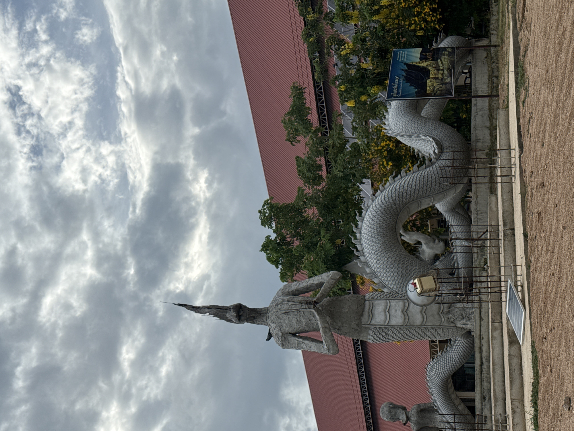 The image depicts a large, unfinished stone sculpture of a dragon or serpent-like mythical creature in a frontal, praying pose. The sculpture is a light gray stone, with detailed scales along its body and a spiky crest on its head. A small bird is perched on the left shoulder of the sculpture. Behind the sculpture, a large building with a distinctive red roof is visible, partially obscured by lush green trees, some of which are bearing yellow flowers. In the foreground, there are construction elements including metal bars and a solar panel on the ground. A sign with Thai script and an image of a temple is mounted on a post to the right of the sculpture. The sky is overcast with dramatic, grey clouds. This scene is located in the city of Cha-am, Thailand.