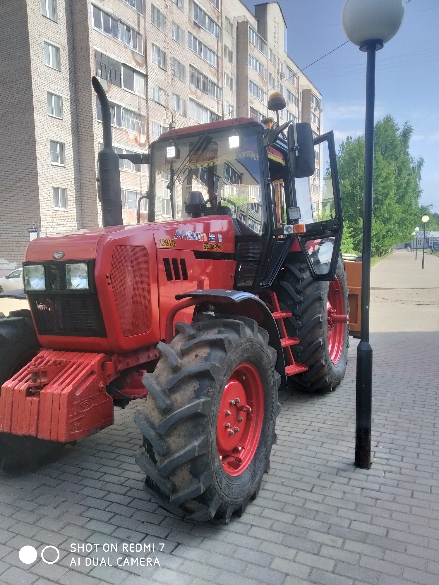 A red Belarus tractor model 1221.3 is parked on a gray brick sidewalk in what is likely the city of Chaikovsky, Russia, based on the tractor's branding in Cyrillic script. The tractor is positioned near a black lamppost with a white spherical light fixture. A multi-story brick apartment building is visible in the background, suggesting an urban residential area. Trees and additional lampposts line the street leading into the distance. No people are clearly visible in the immediate vicinity of the tractor, though there is at least one person visible in the distant background. A car is also parked in the background. The scene appears to be captured on a sunny day. The "SHOT ON REDMI 7 AI DUAL CAMERA" watermark is visible at the bottom of the image.