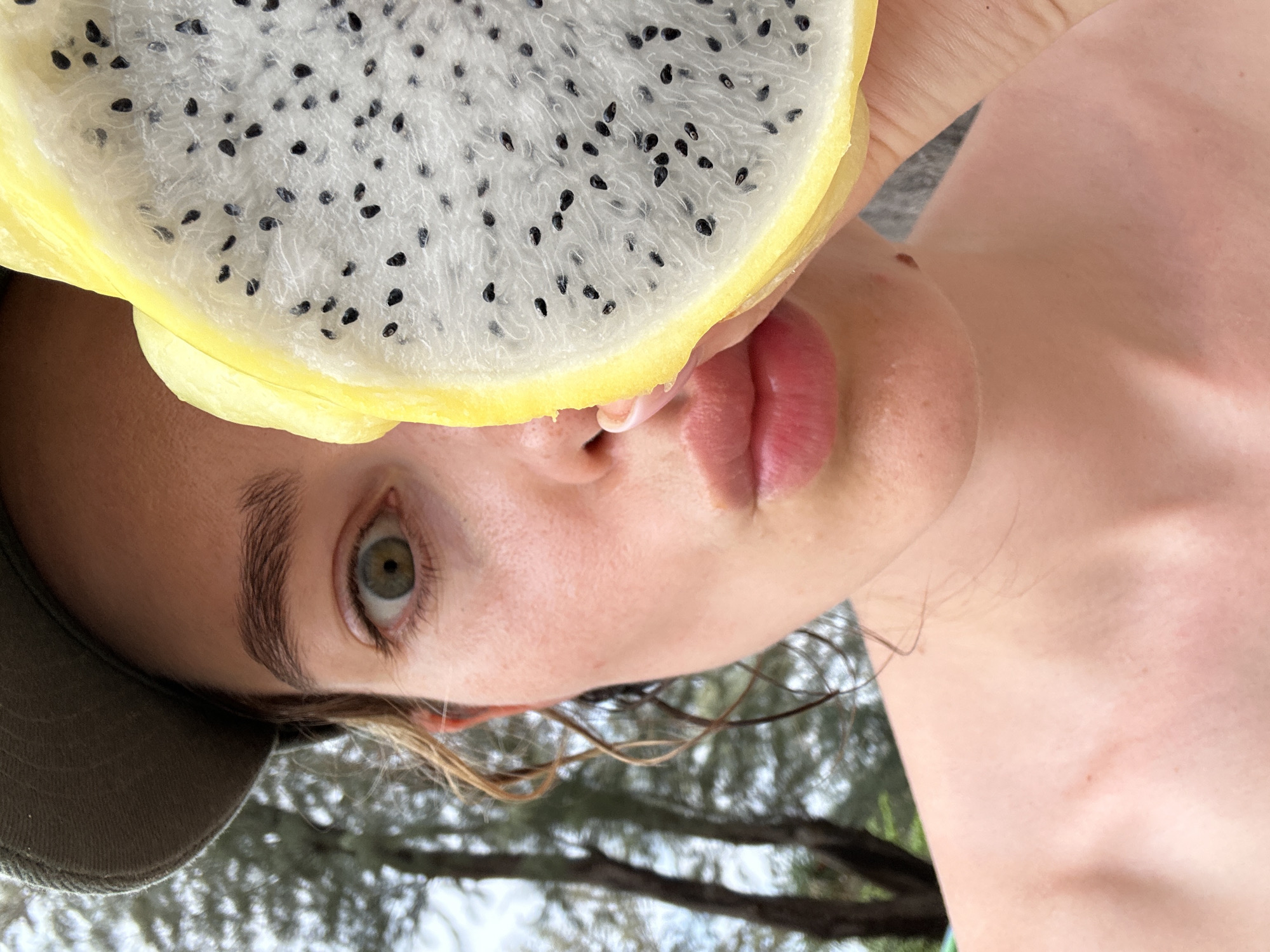 A person is holding a sliced yellow dragon fruit in front of their face. The person is wearing a cap. The person's skin tone is light. The person has an eye color that's mixed green and yellow. The setting is outdoors, likely a natural environment given the presence of trees. The location is Choeng Thale, Thailand.