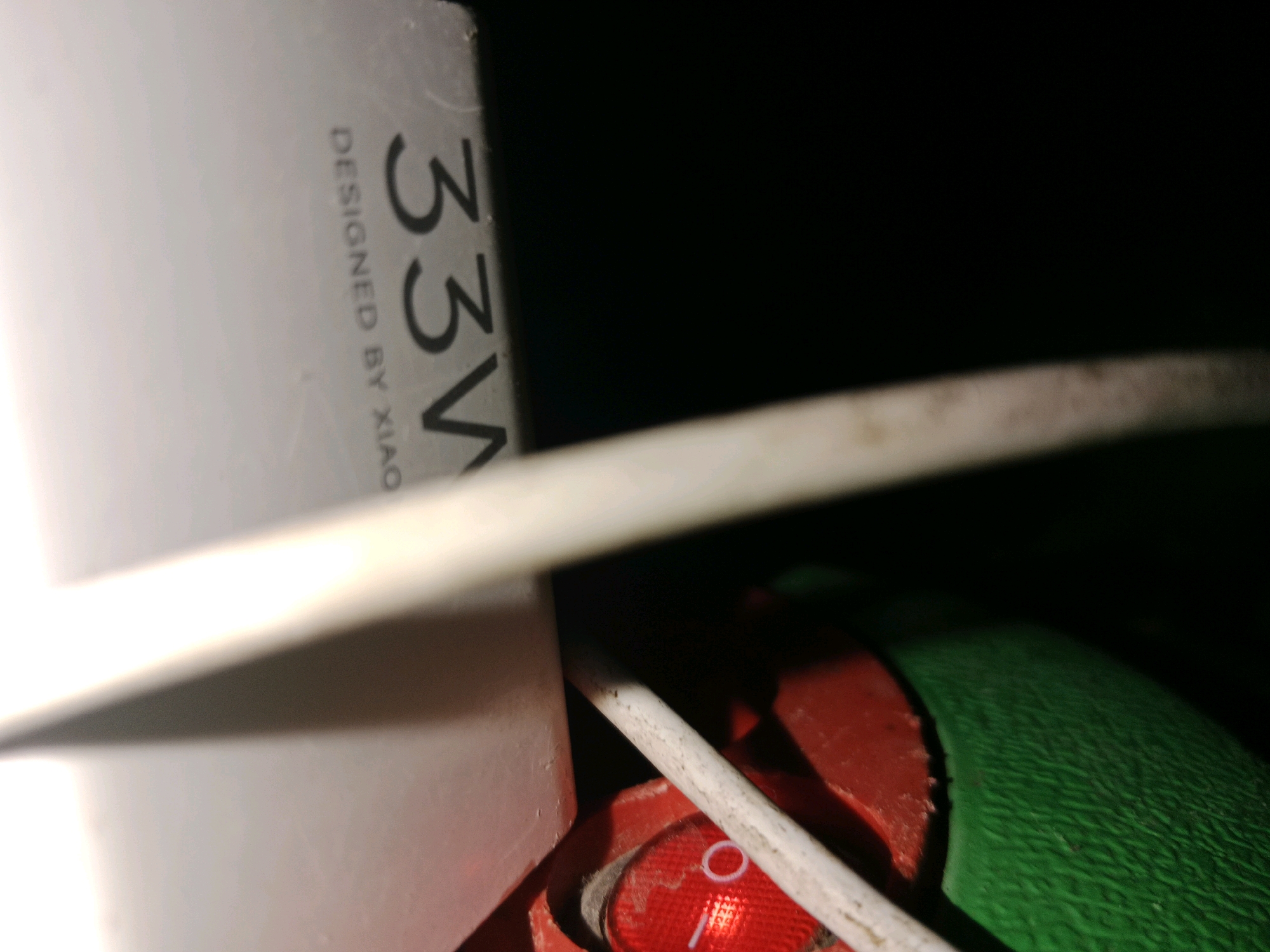 The image displays a close-up, low-angle view of electronic charging equipment against a dark background. A white power adapter, partially visible on the left, has the text "DESIGNED BY XIAO" and "33W" printed vertically on it. A white charging cable extends diagonally across the frame from the top left towards the bottom right, obscuring parts of the adapter and a red power switch. The red switch, located in the bottom center, has a white circular indicator. To the right of the switch, a textured green surface, possibly part of the charging device or a case, is visible. The lighting is dim, highlighting the illuminated red switch. The scene appears to be indoors, with no specific urban or environmental details suggesting a location in Ringinrejo, Indonesia.