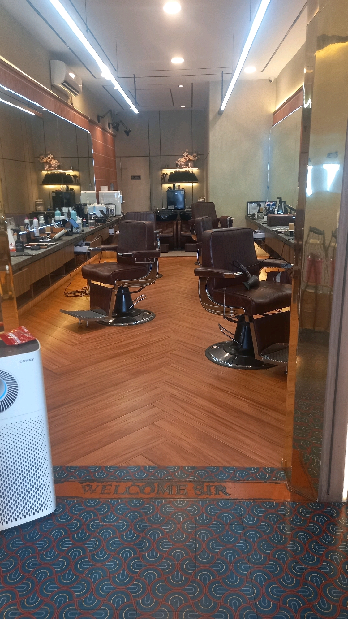 This is an indoor, eye-level, wide shot of a barber shop in the city of Medan, Indonesia. The scene depicts a well-lit, modern establishment with dark brown barber chairs situated on a herringbone-patterned wooden floor. In the foreground, a white air purifier is visible on the left. The backdrop features long counters with various barber supplies, large mirrors, and decorative elements. The overall ambiance suggests a professional and upscale grooming environment.