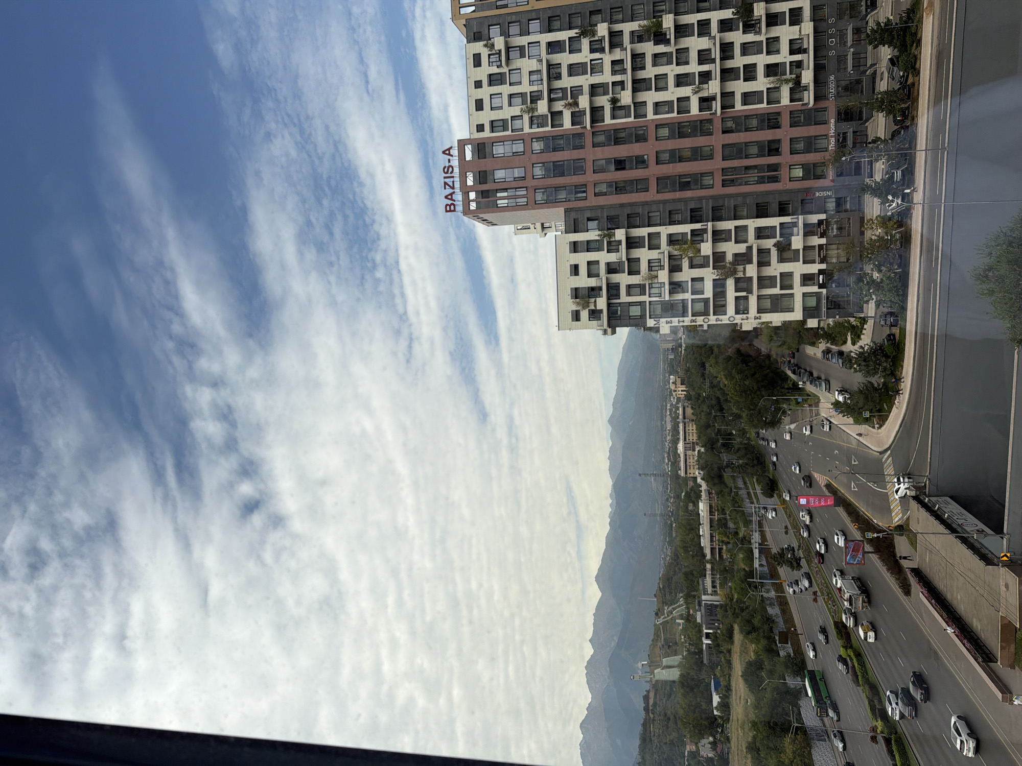 An eye-level, wide-angle shot captures a cityscape with a prominent highway, modern residential buildings, and distant mountains under a partly cloudy sky. The highway, with multiple lanes, is busy with traffic including cars, trucks, and a green bus. Pedestrians are visible on sidewalks and crossing streets. To the right, a cluster of contemporary multi-story apartment buildings with balconies and large windows dominates the mid-ground, with "BAZIS-A" prominently displayed on one building. In the background, a ski jump structure is visible on a tree-covered hill, and beyond that, a range of majestic mountains forms the horizon. The overall scene depicts an urban environment with natural elements integrated into the landscape.