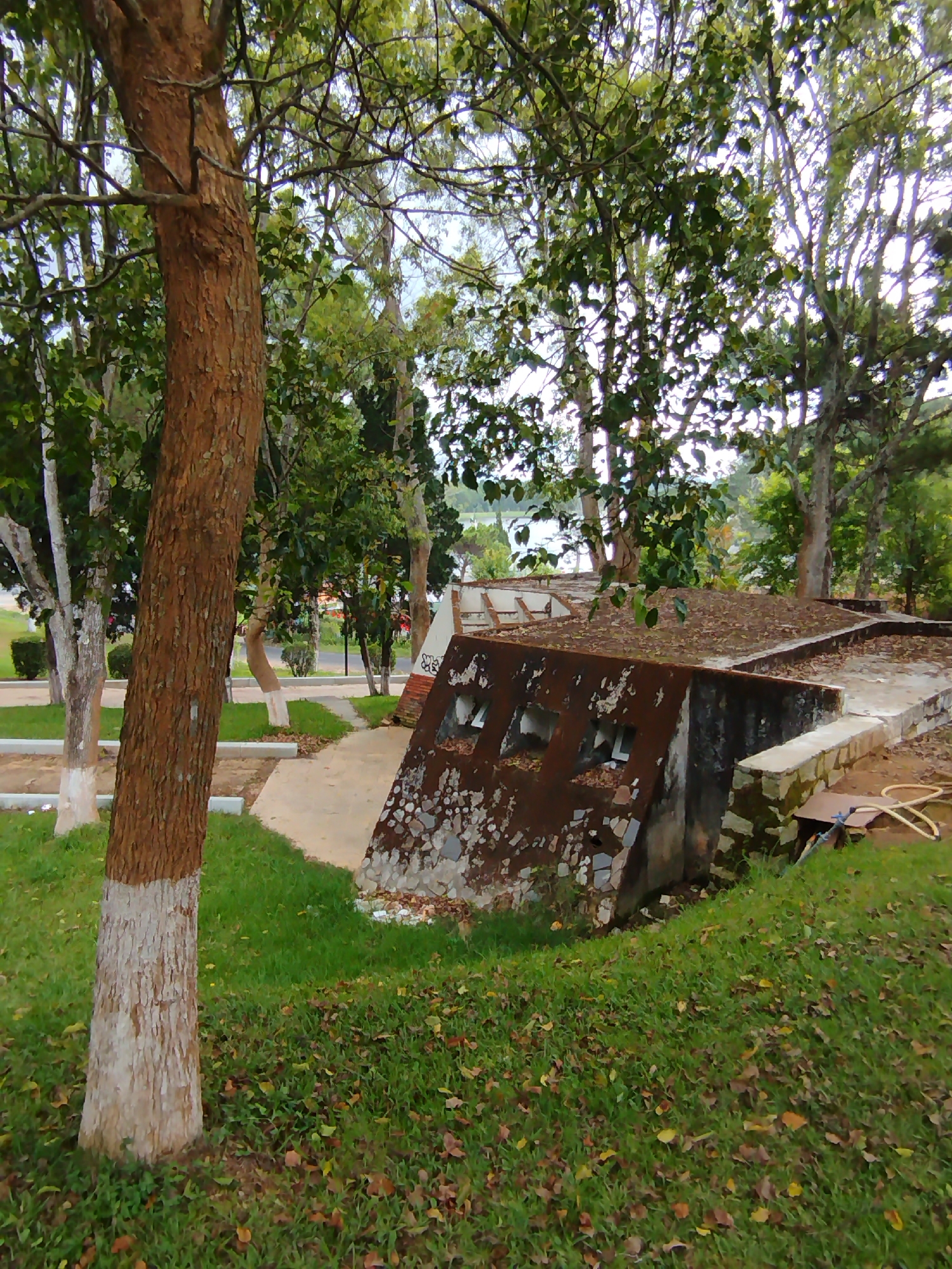 The image shows a partially sunken concrete structure, possibly a bunker or storage facility, located in what appears to be a park in Da Lat, Vietnam. The structure has a rusted exterior and several square openings along one side. It is partly covered with leaves and debris.
To the left, a tree trunk with a light-colored lower portion is visible. Beyond the structure, there is a paved walkway, trees, and grass. In the background, a body of water can be seen. No people are visible in the image. The scene suggests a quiet, somewhat neglected area within the city.