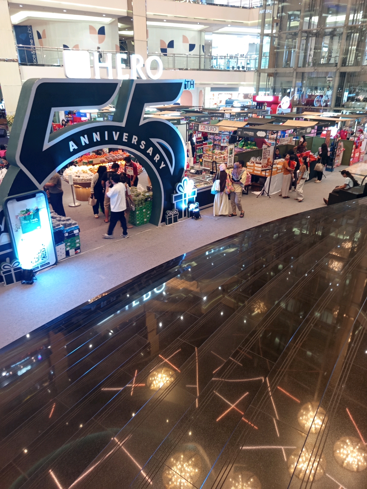 The image shows a large indoor shopping mall, possibly a department store like HERO, celebrating its 75th anniversary. A prominent display features the number "75" in large, stylized green and white lettering, with "ANNIVERSARY" arching around it. Beneath this display, a fresh produce section is visible with various fruits and vegetables. A large, illuminated smartphone screen is also present, displaying the words "Think Fresh." People are seen browsing the produce and surrounding stalls. In the background, multiple levels of the mall are visible, with shops and glass elevators. The floor in the foreground is dark and reflective, with geometric patterns of light. The overall scene suggests a promotional event or ongoing sales within the mall. The location is identified as West Jakarta, Indonesia.