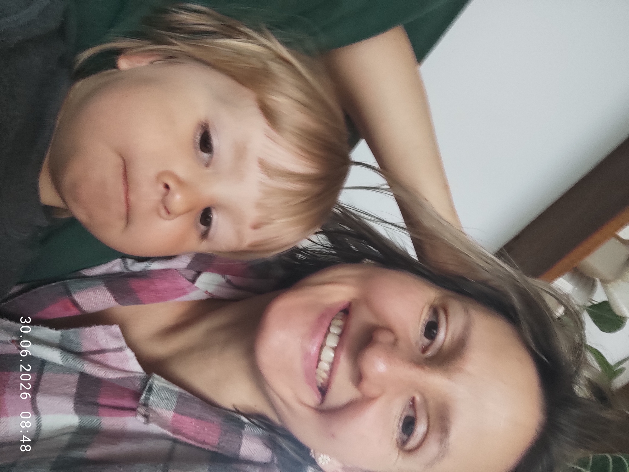 A woman and a young child are captured in a close-up, outdoor, selfie-style photograph. The woman, with shoulder-length brown hair, is smiling broadly and looking towards the camera. She is wearing a pink and grey plaid shirt. To her left, a young child with blonde hair and bangs is positioned, also looking towards the camera with a slightly pouty expression. The child is wearing a dark grey shirt. The background is blurred, but appears to be an indoor setting with a white wall and some plants on a shelf. The timestamp "30.06.2026 08:48" is visible in the lower right corner of the image.