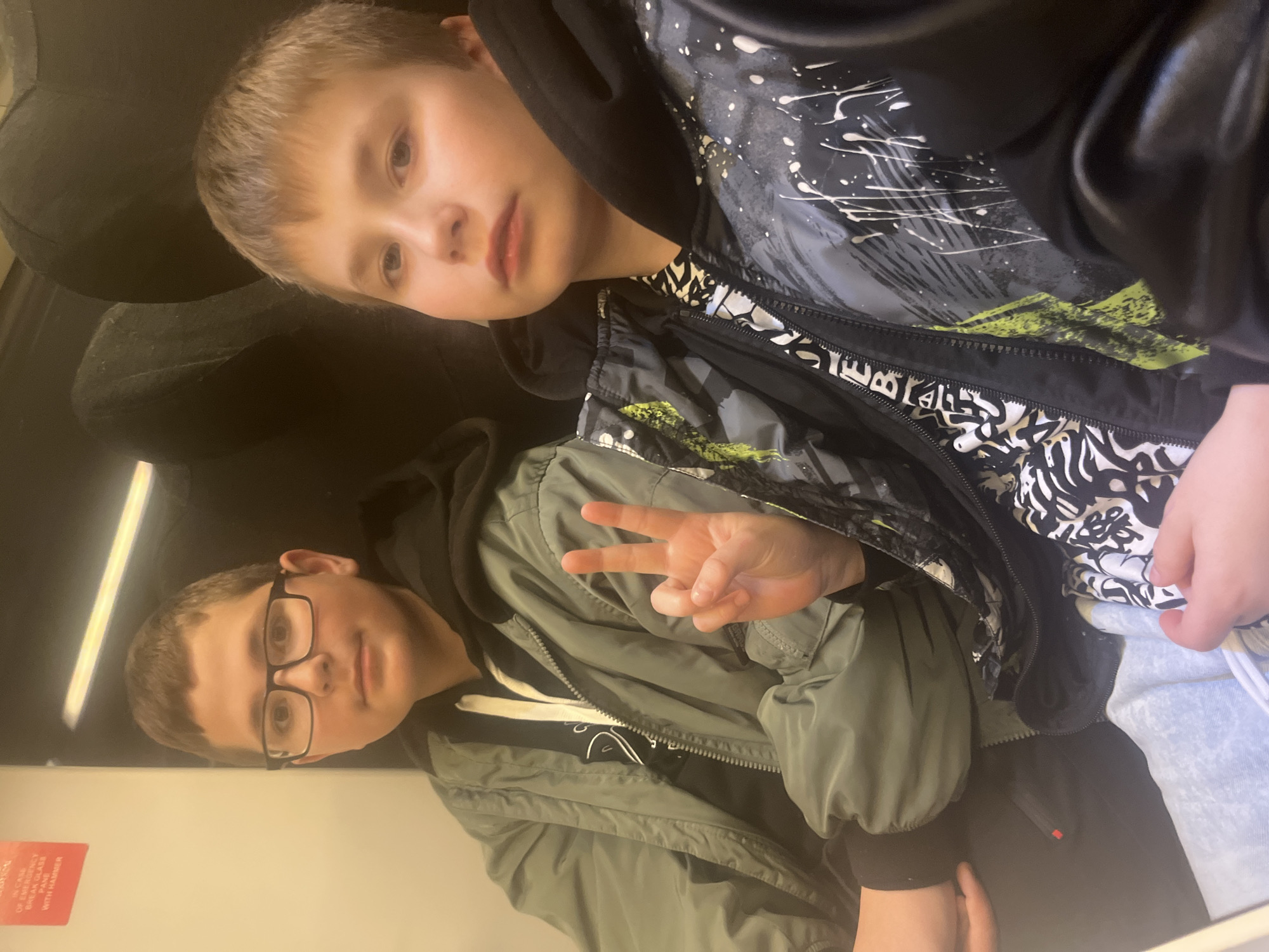 Two Caucasian boys are posing for a selfie indoors, with the image oriented sideways. The boy on the right has short blonde hair, fair skin, and is wearing a black hooded sweatshirt under a patterned jacket. The boy on the left is wearing glasses and a green jacket over a black shirt, and is making a peace sign with his hand. A red emergency sign is visible on the wall behind the boys. The setting appears to be a train or bus. The location is not evident from the image. No information suggests it is in the city of Sochi, Russia.