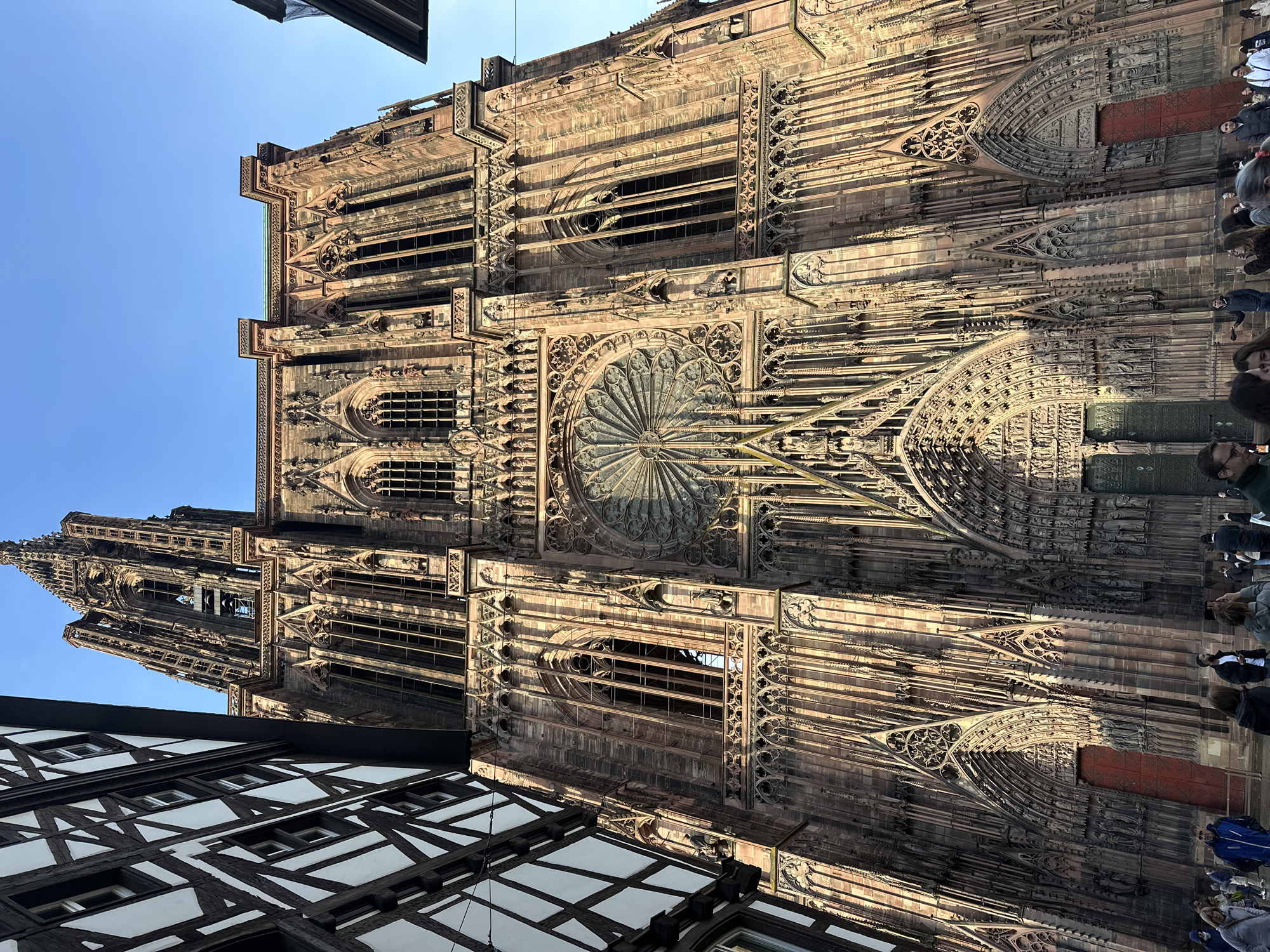 The image shows a portion of the Strasbourg Cathedral, a large Gothic church located in Strasbourg, France. The camera angle is looking up at the building, which is constructed from sandstone. Ornate Gothic architecture is visible, including a large rose window and tall, narrow windows. A section of a building with a traditional timber frame can be seen at the bottom left of the image. People are standing at the base of the cathedral on the right-hand side. The sky is light blue.