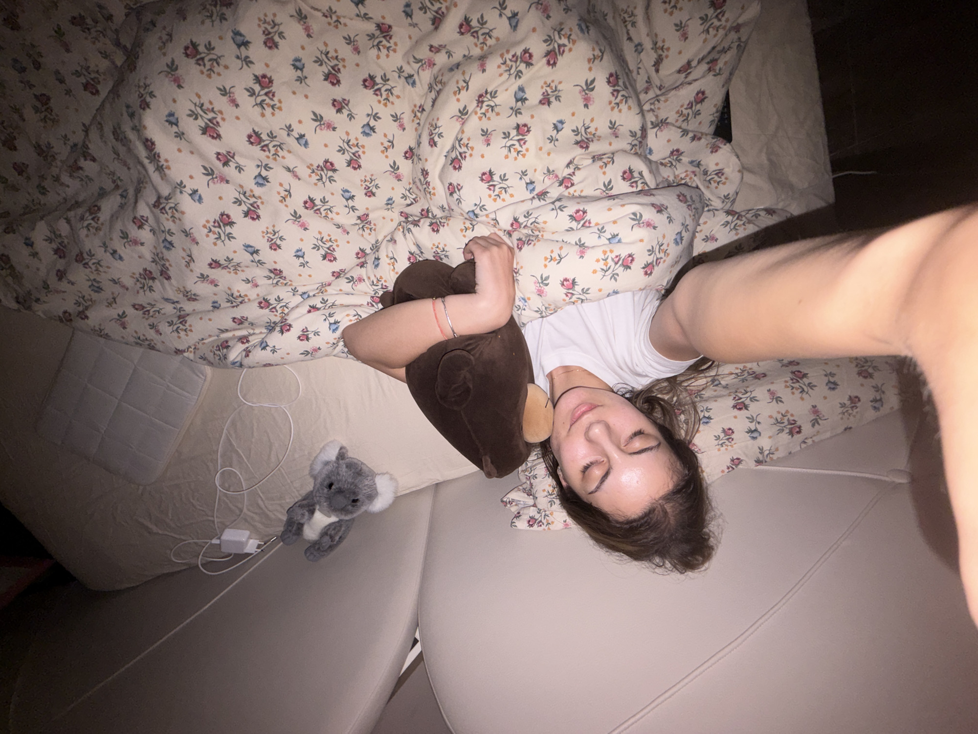 A young woman lies in a bed covered with a floral patterned blanket. She is holding a brown stuffed animal. A gray koala stuffed animal and a white phone charger are on the bed next to her. The room is dimly lit. There is no indication of this image's relationship to the city of Burgas, Bulgaria.