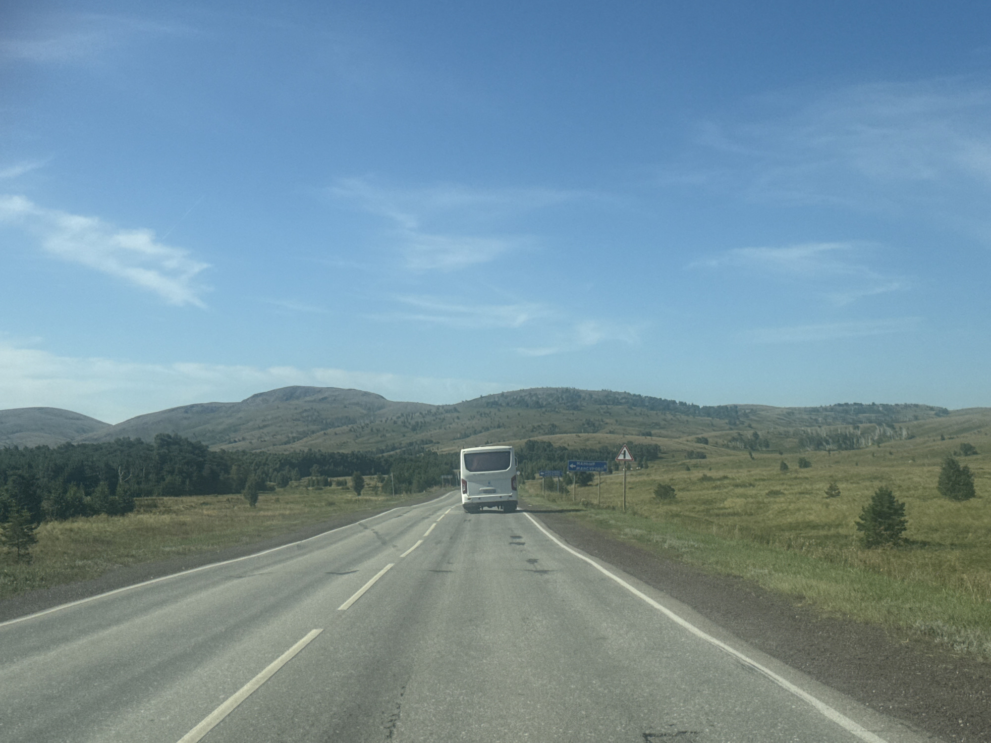 An asphalt road stretches into the distance, leading through a rural landscape under a clear blue sky with wispy clouds. The road is marked with white lines, and a white bus is visible in the center of the frame, traveling away from the viewer. On either side of the road, there are grassy fields with scattered trees and rolling hills covered in vegetation. In the distance, larger hills form the horizon. A road sign with a warning symbol and directional signs are posted on the right side of the road. The scene appears to be a view from a moving vehicle, likely captured on a sunny day. The location is identified as being near the city of Baymak, Russia.