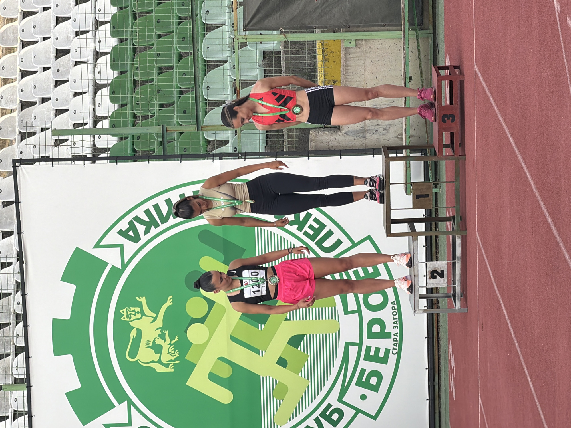The image displays an awards ceremony for a track and field event, featuring three female athletes on a multi-tiered podium. The scene is set outdoors at a stadium in Stara Zagora, Bulgaria, with a red running track in the foreground and stadium seating visible in the background.
The three athletes stand on a wooden podium, with positions clearly marked. The first-place winner (center, highest tier) wears a black athletic top with a bib displaying "1200", pink shorts, and light-colored shoes, proudly showing a gold medal. The second-place athlete (right, middle tier) is dressed in a light brown top, black leggings, and black and pink athletic shoes, wearing a silver medal. The third-place athlete (left, lowest tier) wears a red athletic top, black shorts with white stripes, and pink athletic shoes, with a bronze medal around her neck. The numbers "2" and "3" are visible on their respective podium steps.
Behind the podium is a large white and green banner. It features a stylized green logo with a running figure and an animal resembling a lion or griffin. Visible Cyrillic text on the banner includes "ЛЕКА АТЛЕТИКА" (Light Athletics/Track and Field) at the top, "КЛУБ 'БЕРО'" (Club 'Bero') at the bottom, and "СТАРА ЗАГОРА" (Stara Zagora) along the bottom curve, confirming the event's location.
The background shows stadium seating with a mix of white and green plastic chairs. The lighting suggests it is daytime, possibly bright but overcast, as no harsh shadows are evident. The athletes appear composed and content, likely posing for a commemorative photograph after their successful competition.