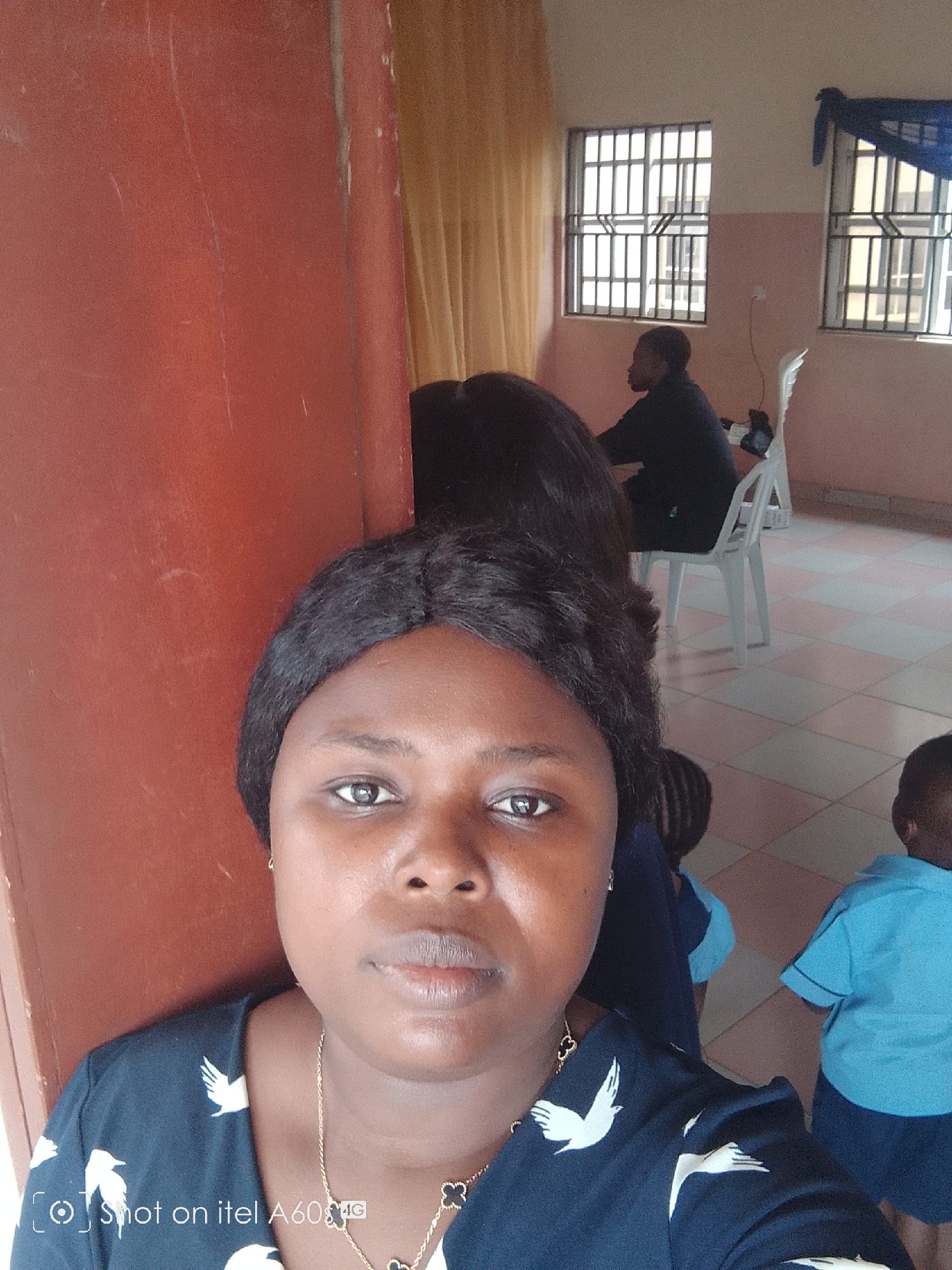 The image depicts a woman taking a selfie, with a red wall on her left. In the background is a classroom with two barred windows, a woman sitting on a white chair, and two children wearing blue uniforms. The location is implied by the camera watermark as being in Idimu, Nigeria, due to it being shot on an Itel A60 smartphone.