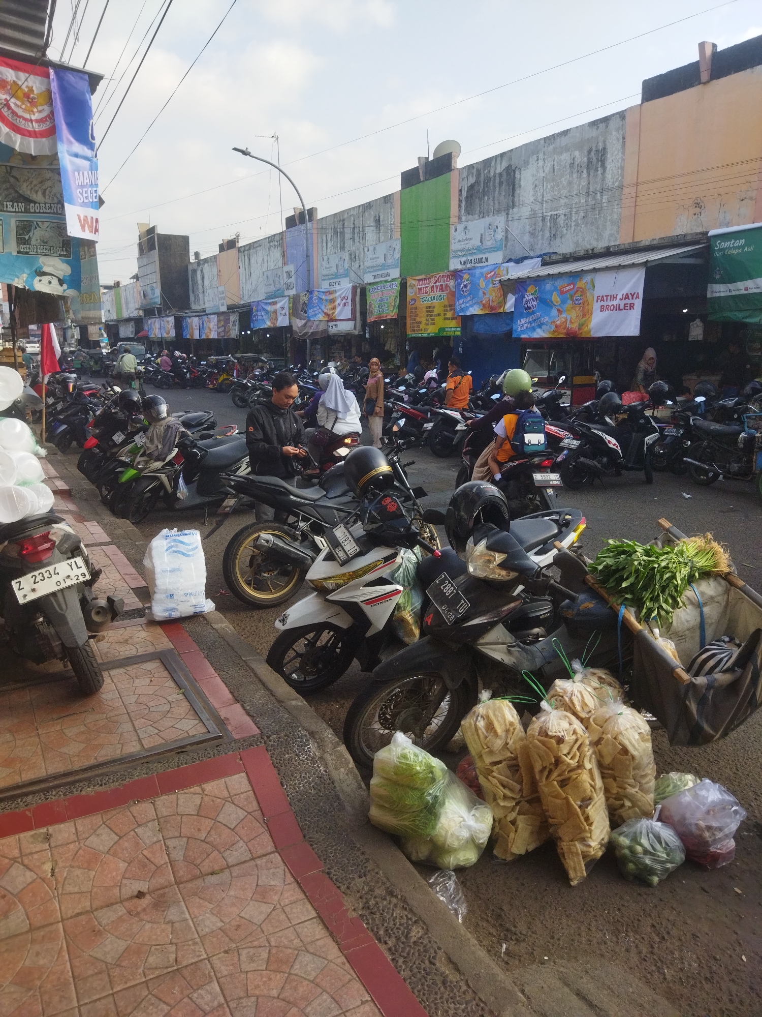 The image depicts a street scene in Banjar, Indonesia, likely a market area. In the foreground, several motorcycles are parked along a tiled sidewalk. A man in a black jacket is standing by a motorcycle, holding a mobile phone. Bags of groceries, including vegetables and packaged snacks, are placed on the ground near the motorcycles.
The background shows a row of buildings with colorful banners advertising various products. The buildings appear to be shops or stalls, with people milling about in front of them. The sky is visible at the top of the image, with some clouds. The overall impression is of a busy, bustling commercial area in an Indonesian city.