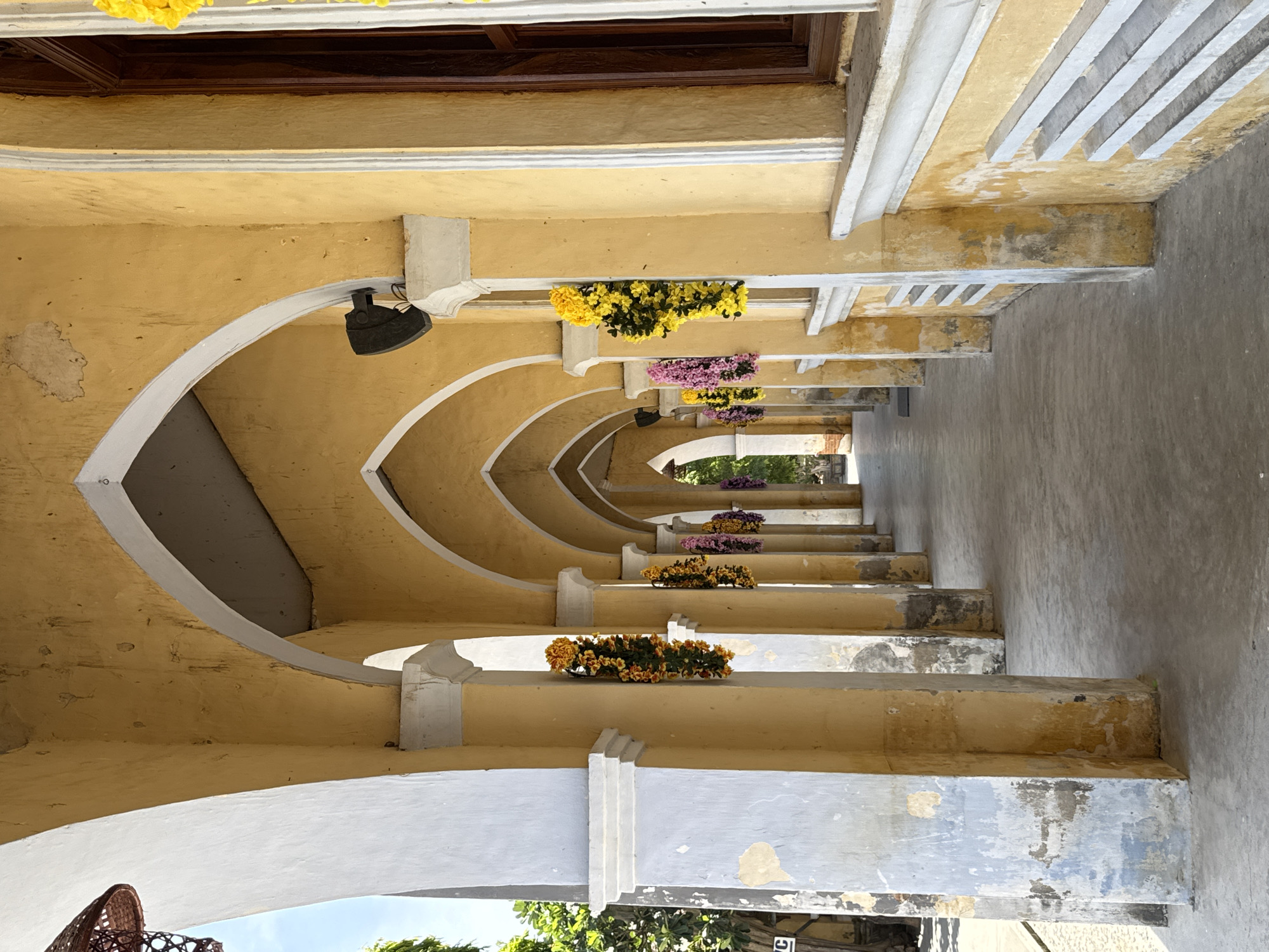 This is a photograph taken outdoors, likely during the daytime given the bright lighting and shadows. The scene depicts an outdoor walkway with a series of arched openings leading into the distance. The architecture features a predominantly yellow-colored facade with white trim along the arches and pillars. Several planters with yellow and pink flowers are arranged along the walkway, adding pops of color to the aged, somewhat weathered structure.
The walkway appears to be a covered area, possibly part of a historical building or a temple complex given the architectural style and the presence of potted flowers which are common decorative elements in such settings in Vietnam. The ground is a paved walkway, showing signs of wear. The light suggests it's a sunny day. There are no people visible in the frame. The perspective is from a low angle looking down the length of the walkway, creating a sense of depth. The overall impression is one of a tranquil, perhaps historical, outdoor space decorated with simple floral arrangements.