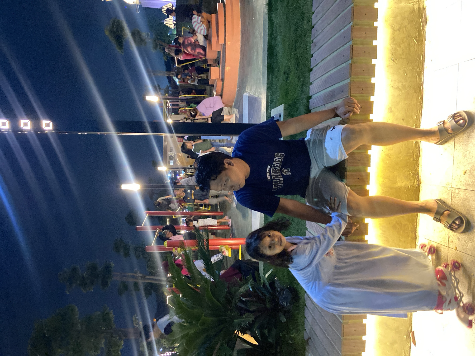 This media file depicts a nighttime scene at an outdoor venue, possibly an amusement park or a public space with recreational facilities. Several people are visible in the background, some appearing to be seated on benches or structures. In the foreground, a man wearing a dark blue t-shirt with "NEW YORK YANKEES" and the team's logo in white, along with light-colored shorts, is standing. Next to him, a young girl in a light blue outfit is also present, facing the camera and smiling. She appears to be wearing roller skates. The setting is illuminated by artificial lights, including string lights and ground lighting, creating a vibrant atmosphere. Some areas are grassy, and wooden structures are also visible. The sky is dark, indicating it is nighttime. The overall mood seems relaxed and enjoyable, with the subjects appearing to be in good spirits. There are also trees and foliage present, suggesting an outdoor environment. The location is identified as Sidoarjo, Indonesia.