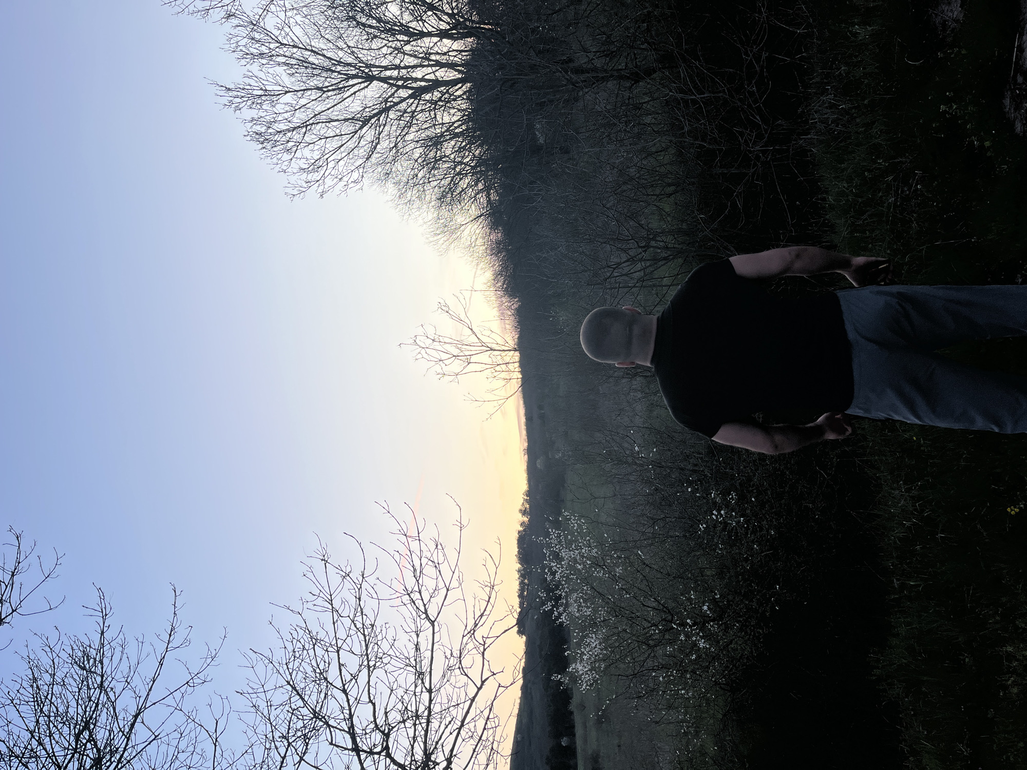 A man stands in a field at dusk, his back to the camera. He is wearing a black t-shirt and blue jeans. The sky is a pale blue, with a hint of orange on the horizon. There are trees in the background, some with bare branches and others with small white flowers. The ground is covered in grass and dark vegetation. Location appears to be a rural area near Burgas, Bulgaria.