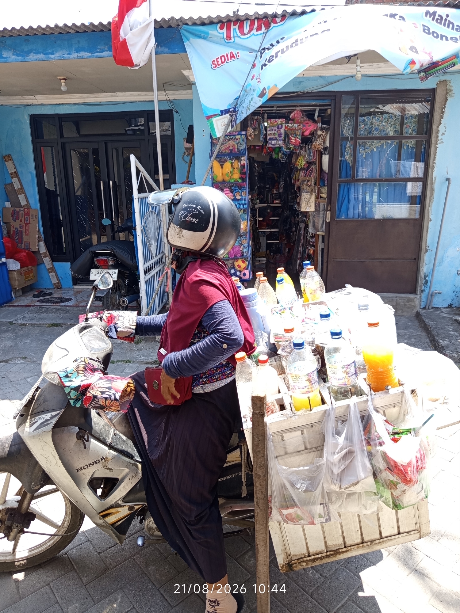 In the city of Canggu, Indonesia, a person wearing a red headscarf and dark clothing is seated on a scooter. They are wearing a black and white helmet with the words "Freedom Rider" and "Classic" printed on it. The scooter is parked on a brick-paved street in front of a small shop. The shop has a blue facade and a large banner above the entrance displaying text in Indonesian. The shop appears to sell a variety of goods, including drinks in plastic bottles and packaged snacks. A wooden cart filled with more drinks and plastic bags of goods is attached to the side of the scooter. The scene is illuminated by bright daylight, casting shadows on the ground.