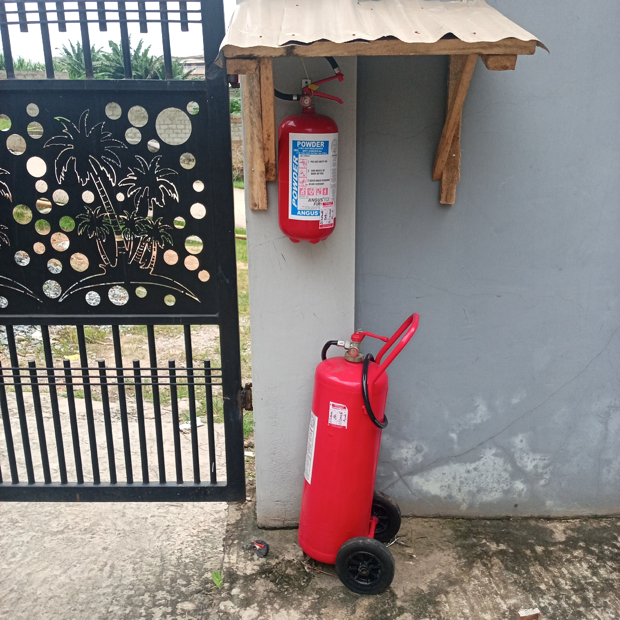 In the city of Ibadan, Nigeria, a scene unfolds in front of a grey concrete wall and a black metal gate with a palm tree motif. Two red fire extinguishers are prominently displayed. One, smaller and cylindrical, hangs from a makeshift wooden shelter attached to the wall. This extinguisher has a white label with blue lettering that reads "POWDER" and instructions for use. The second, larger fire extinguisher stands on two black wheels on the ground, leaning against the wall. It also has a white label with red and blue text, and its hose is coiled around its body. The ground in front of the wall and gate is a mixture of concrete and gravel. In the background, through the gate, vegetation and distant buildings are visible.