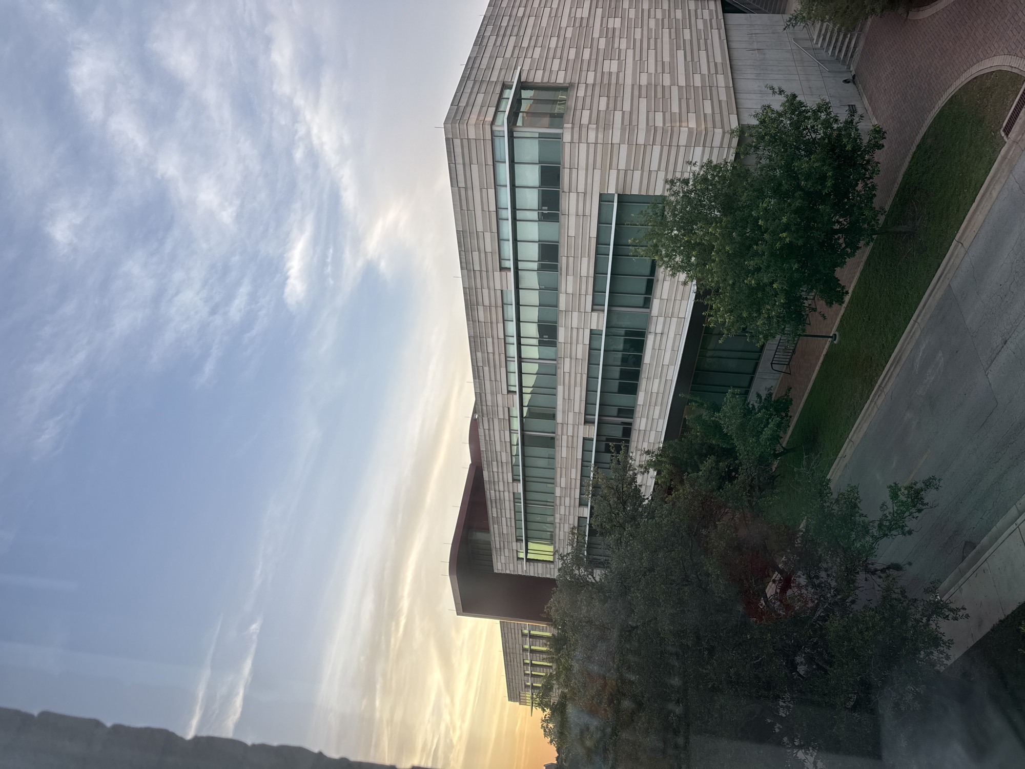 The image depicts a modern building with a beige brick facade and large windows, likely an office or academic building. The building is situated along a street with trees and landscaping. The sky shows a sunset or sunrise, with clouds illuminated by the warm light. The location is likely in Austin, Texas, based on architectural styles and the overall environment. The scene is captured from an elevated position, possibly from another building or a high floor, providing a view over the landscape.