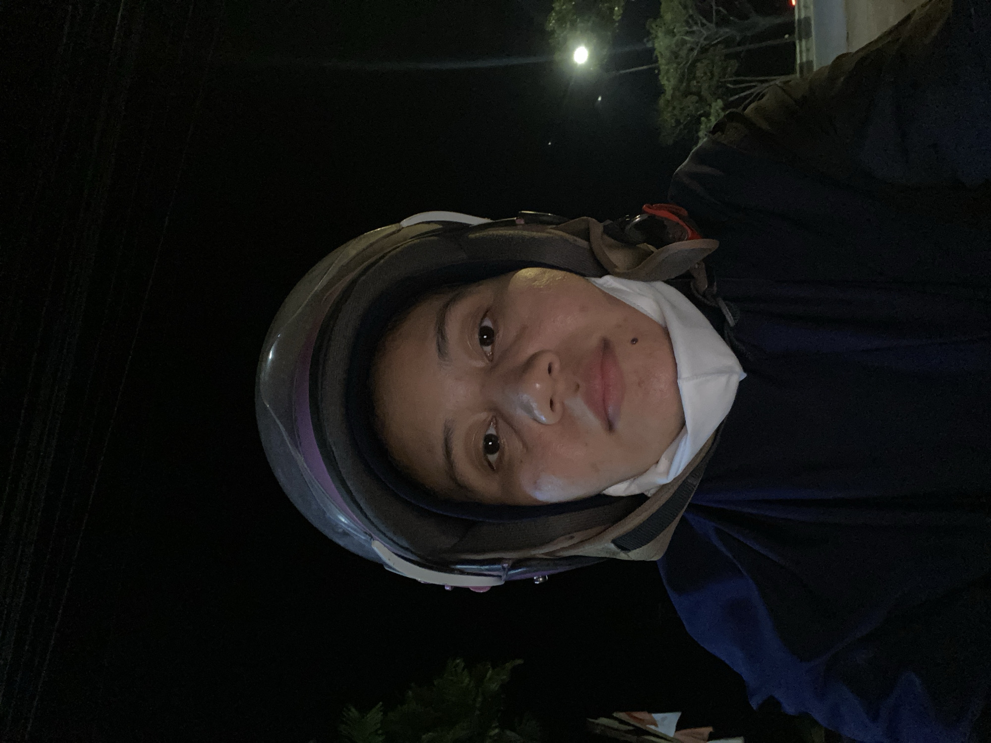 A close-up, front-facing, selfie-style photograph shows a person wearing a dark blue jacket and a helmet in Gambut Barat, Indonesia, at night. The person's face is visible, with eyes open and looking at the camera. A white mask is worn around the neck, covering the chin. The background is dark, with some faint lights and trees visible, suggesting an outdoor setting. The overall lighting is low, with a light source illuminating the person's face from the front.