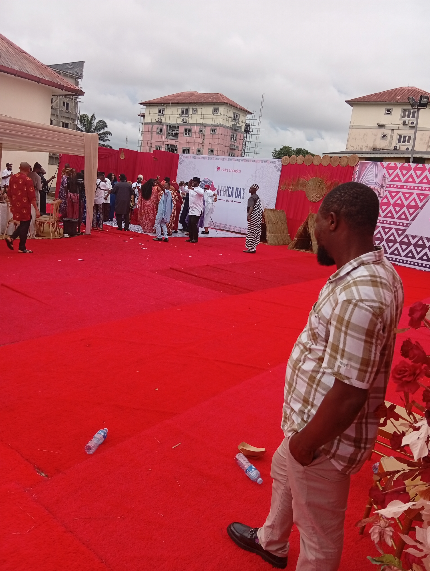The image shows a gathering in Rumuepirikom, Nigeria, possibly an event related to "Africa Day," as indicated by a backdrop sign. A red carpet covers a large area, and several people are present, some dressed in traditional African attire. There's a man in a plaid shirt and tan pants standing with his hand in his pocket in the foreground, facing toward the rest of the crowd. There are also water bottles discarded on the carpet. Buildings are visible in the background, some under construction. The sky appears overcast.