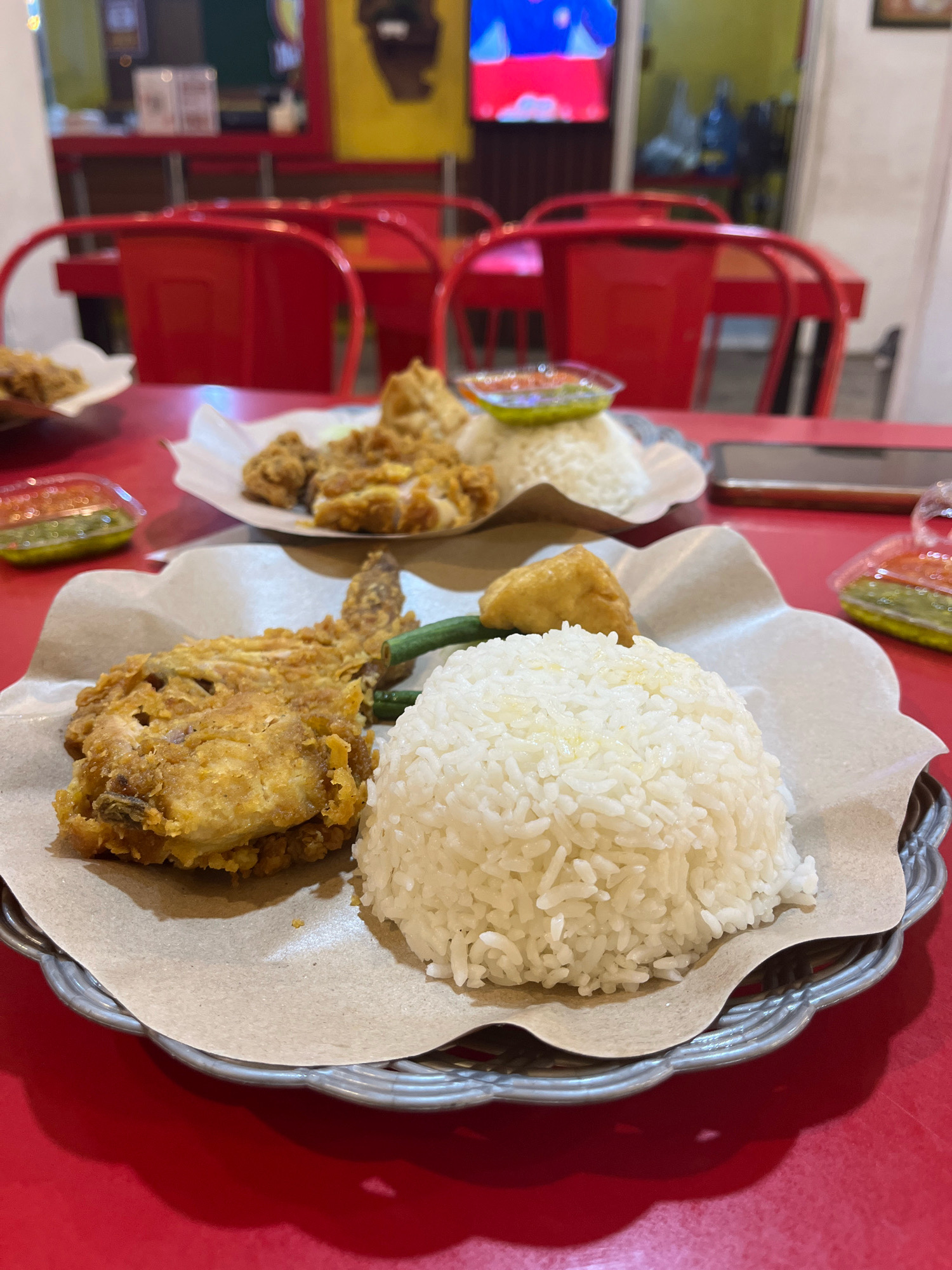 The image shows a close-up of two plates of fried chicken and rice on a red table, likely in a restaurant in Citrodiwangsan, Indonesia. The plate in the foreground features a large piece of crispy fried chicken, a generous portion of white rice, a few green beans, and a piece of fried tofu. A small plastic container with green sauce is visible to the left. The plate in the background, slightly out of focus, also displays fried chicken and rice, with another small container of sauce. Red chairs are visible in the background, suggesting a dining setting. The lighting is warm, and the overall scene suggests a casual meal.