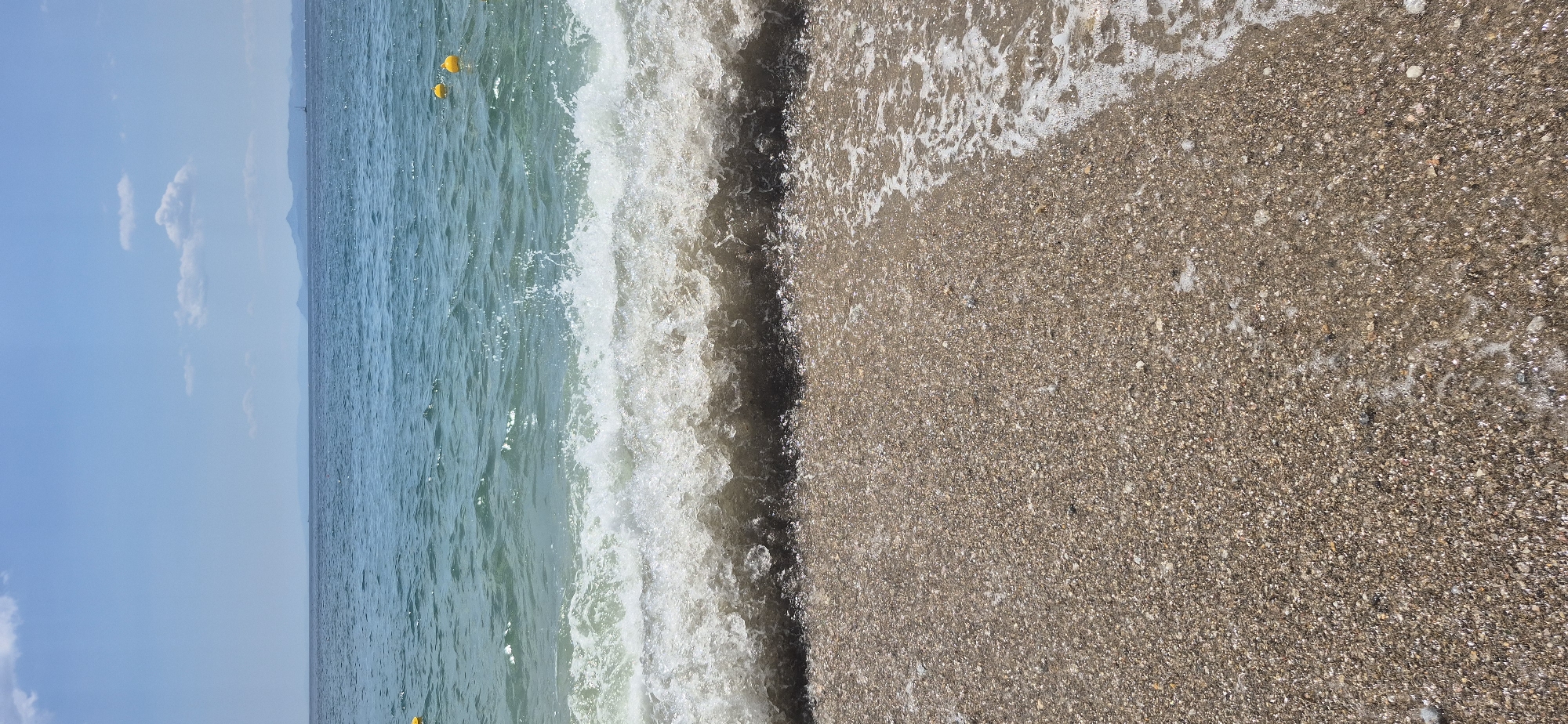 This media file captures a serene beach scene in Paleo Faliro, Greece. The image is oriented vertically, presenting a split view of the sea and the shore.
On the left, the tranquil blue sea stretches towards the horizon, meeting a clear sky dotted with a few wispy white clouds. The water is calm, with gentle ripples reflecting the sunlight. In the distance, faint mountains are visible, hinting at the vastness of the landscape. Two bright yellow buoys float on the surface of the water, suggesting a designated swimming or boating area.
The right side of the image is dominated by the shoreline. A wave is captured in the act of breaking, its white foam contrasting with the deeper blue of the sea. The water washes over the coarse, pebbly sand, creating a wet sheen and leaving a thin line of foam. The sand itself is a mix of light and dark grains, interspersed with small shell fragments, giving it a textured appearance.
The overall impression is one of a peaceful summer day. The bright sunlight indicates it is likely daytime, and the clear sky suggests fair weather. The scene evokes a sense of relaxation and tranquility, characteristic of a Mediterranean beach. There is no visible text in the image.