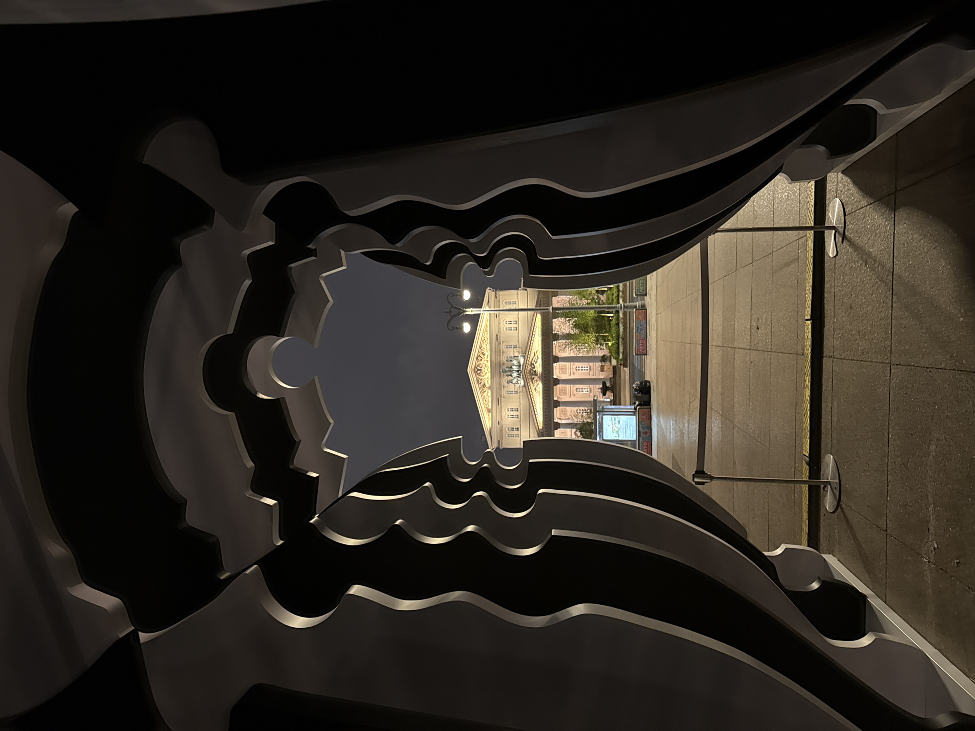 The image depicts a nighttime urban scene in Moscow, Russia, framed by an abstract, black-and-white metal sculpture. The sculpture has several layers with a wavy, cut-out design. Through the center of the sculpture, a view of a street reveals the Bolshoi Theatre, a prominent landmark in Moscow. A street lamp stands in front of the theatre. On the right side of the image, there is a paved sidewalk and some metal poles with discs attached to them. In the background, there is a light box advertising.