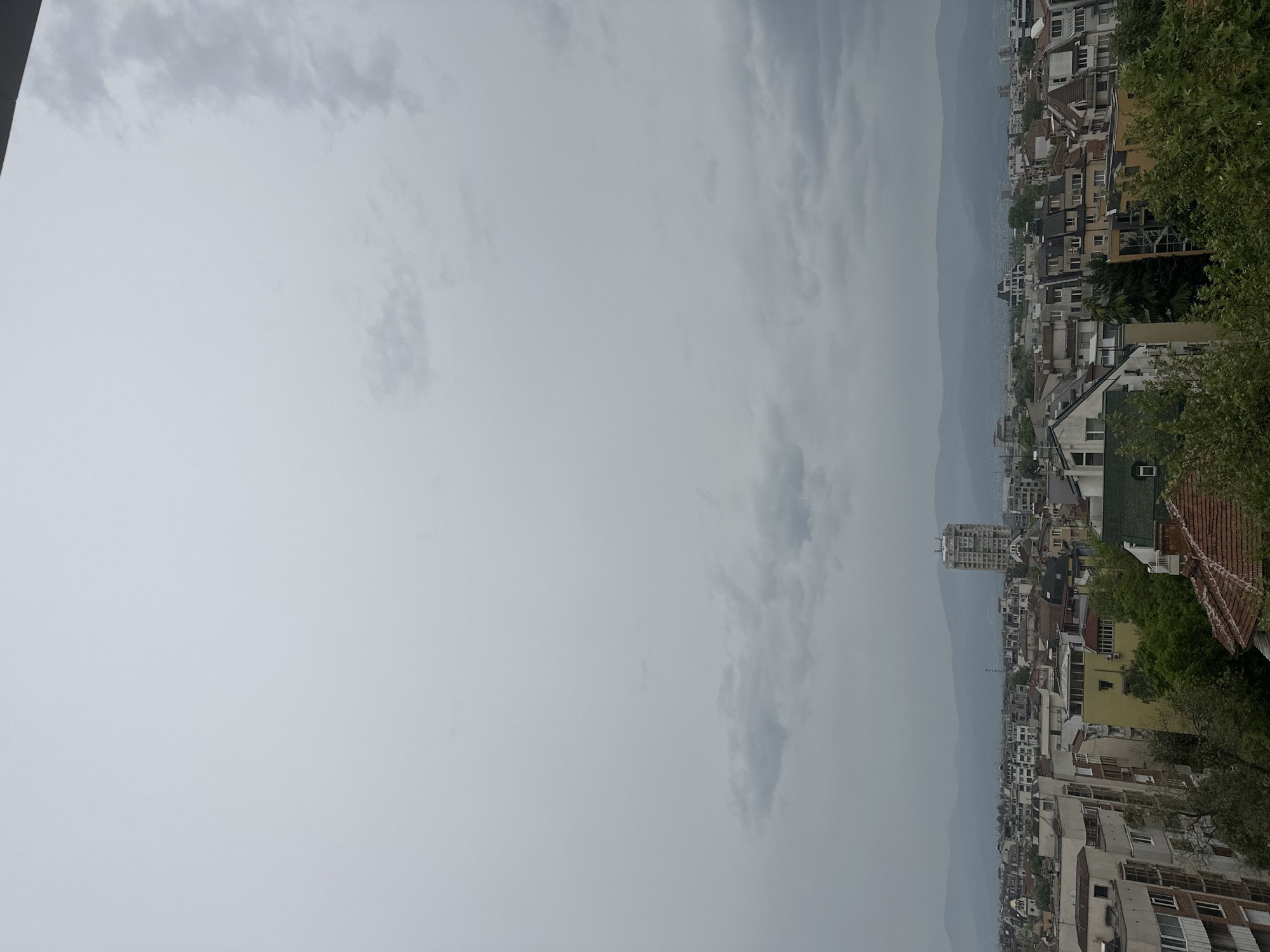 The image depicts a city landscape under a cloudy sky. The city is densely populated with buildings of varying heights and architectural styles, including both low-rise residential structures and a few taller buildings. In the background, a mountain range is visible, partially obscured by haze or clouds. The overall weather condition appears to be overcast or rainy. Given the location is Plovdiv, Bulgaria, it is difficult to determine a precise neighborhood without additional information.