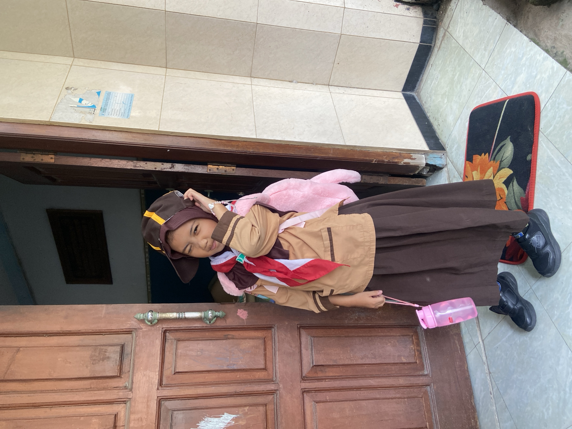 A young girl in a brown and tan scouting uniform, complete with a neckerchief and hat, stands in front of a wooden door. She holds a pink water bottle. The location appears to be the entrance of a home in Gelam, Indonesia, as evidenced by the architectural style and the presence of a welcome mat with a floral design. Tiles cover the porch. The girl is positioned between the opened door and a decorative rug on the porch.