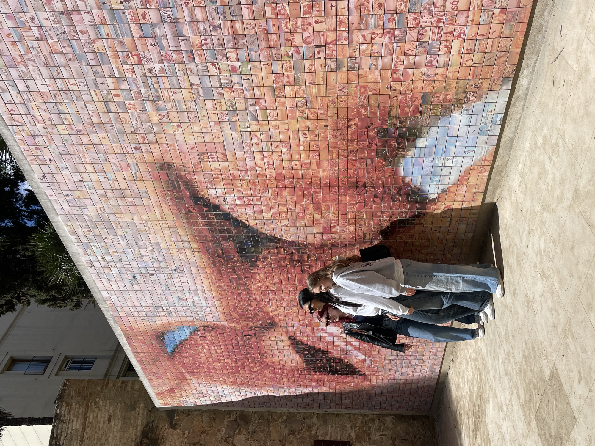 A low-angle outdoor shot depicts three young women standing in front of a large mural composed of numerous small tiles, collectively forming an image of a human face. The mural appears to be affixed to a building or structure, with the tiled face spanning a significant portion of the background. The women are standing close to the mural. The mural is known as "The world begins with every kiss" and is located in Barcelona. The surrounding environment includes a portion of a building visible on the left side and a paved area on the right. The lighting suggests a sunny day.