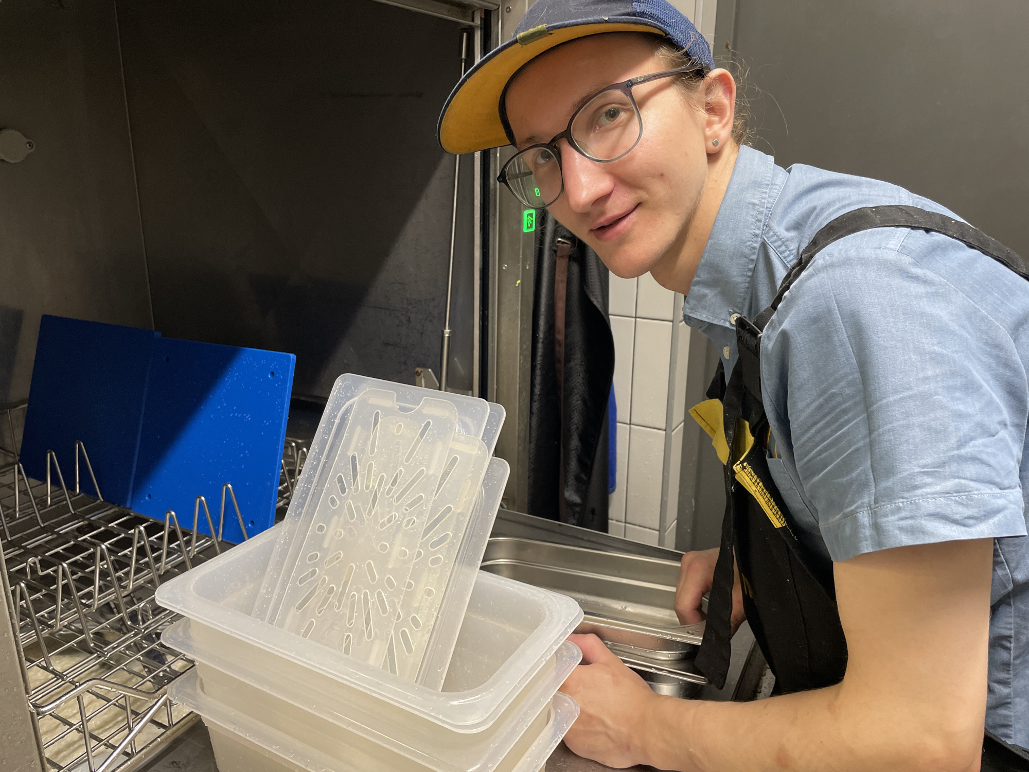 A person wearing a blue shirt, black apron, and a blue and yellow baseball cap is standing in what appears to be a kitchen or food preparation area. They are looking towards the camera with a slight smile. In front of them, there are several clear plastic food containers stacked. To the left, there is a metal rack holding a bright blue cutting board. The environment seems to be indoors, with stainless steel surfaces and tiled walls visible. The person's hands are visible, reaching towards the containers.